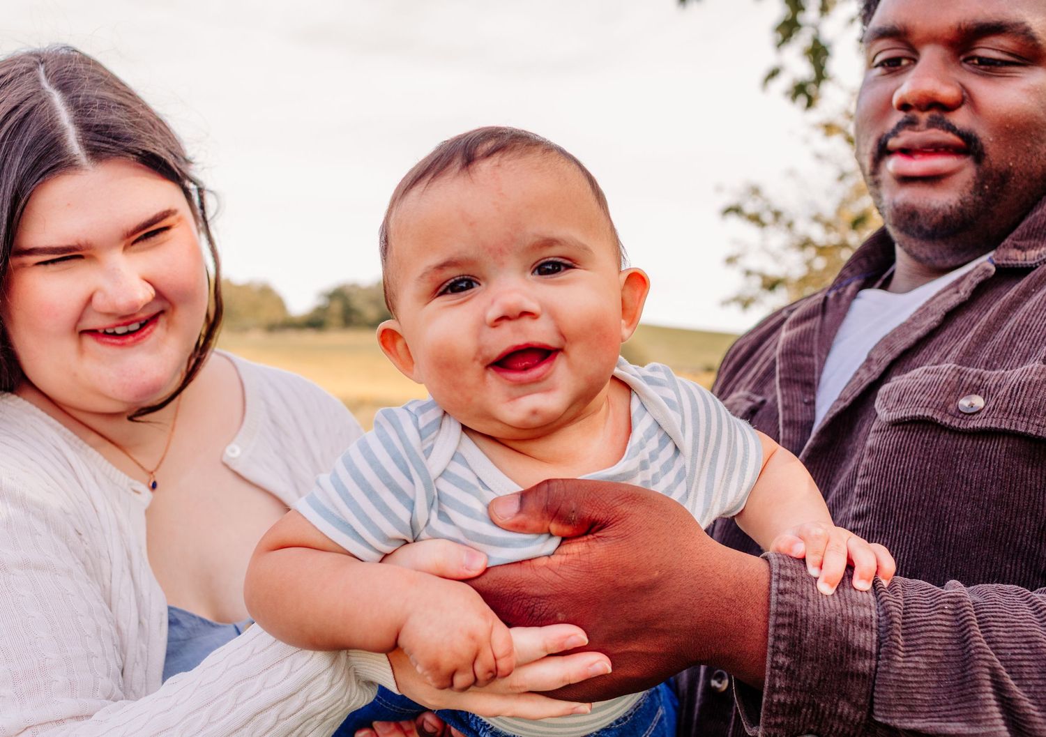 Close-up of joyful baby smiling between loving interracial parents during outdoor family portrait.