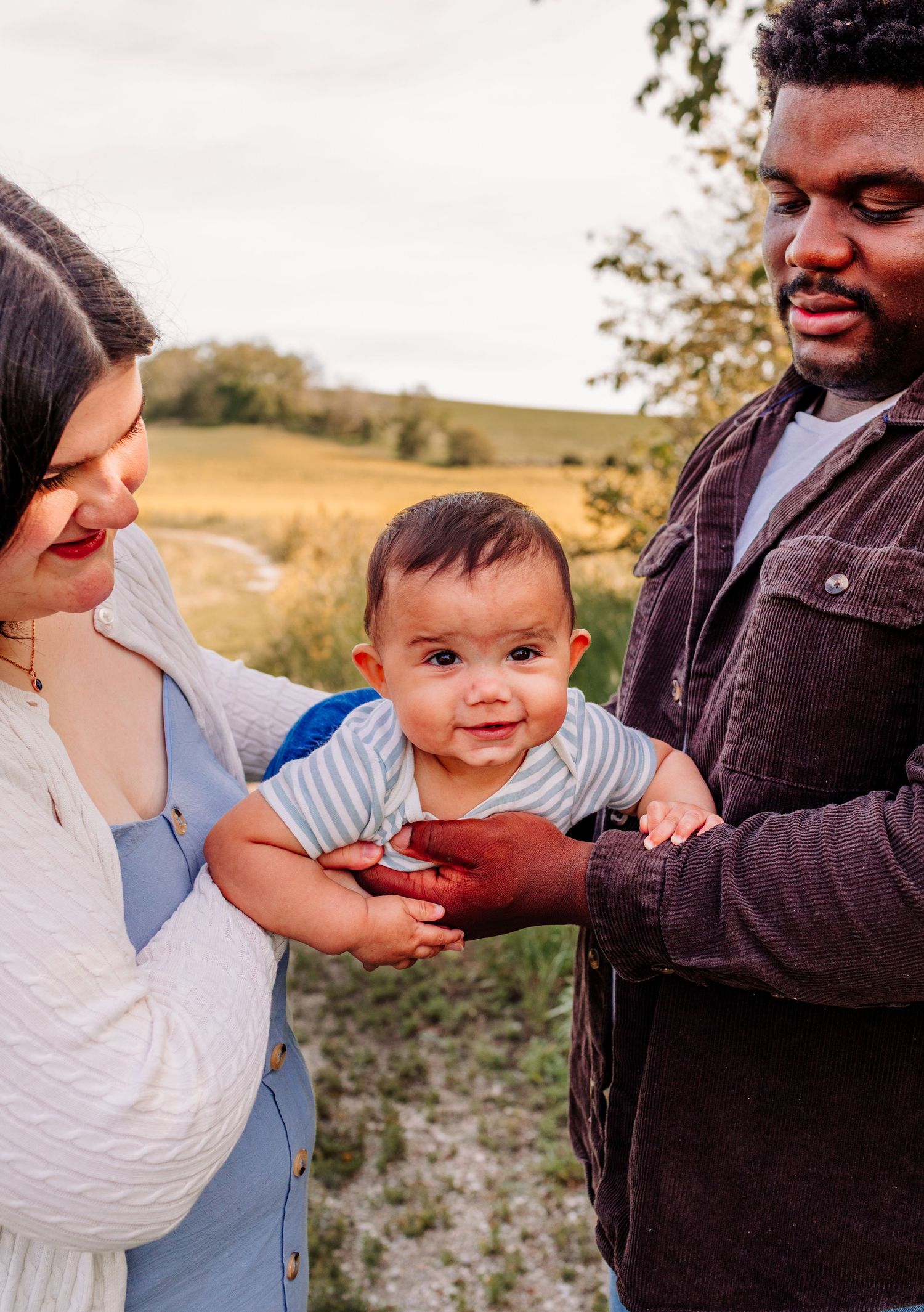 Loving interracial couple holding their smiling baby boy outdoors in a sunlit field during a family photo session.