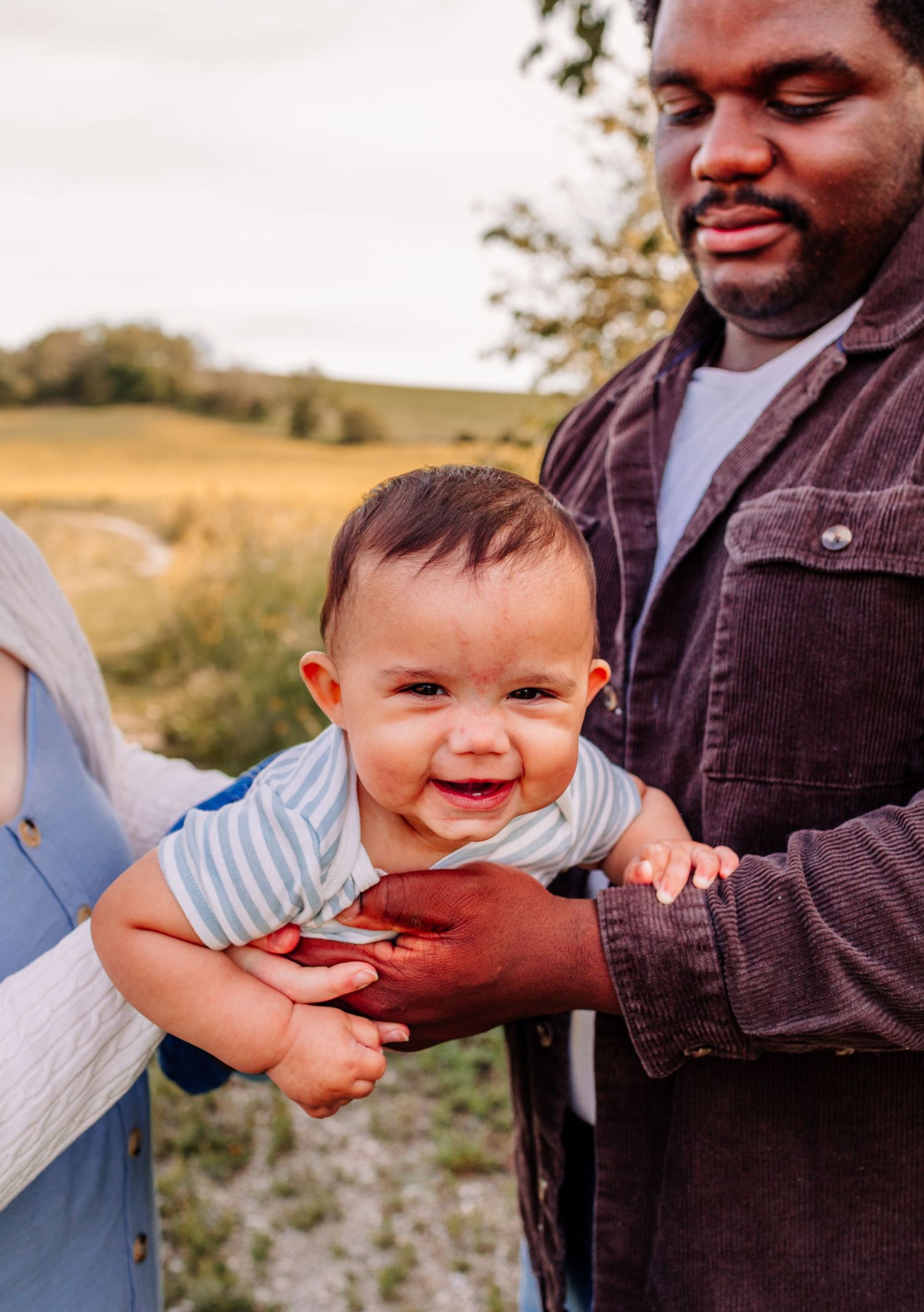 Laughing baby held by father in brown jacket in outdoor field.