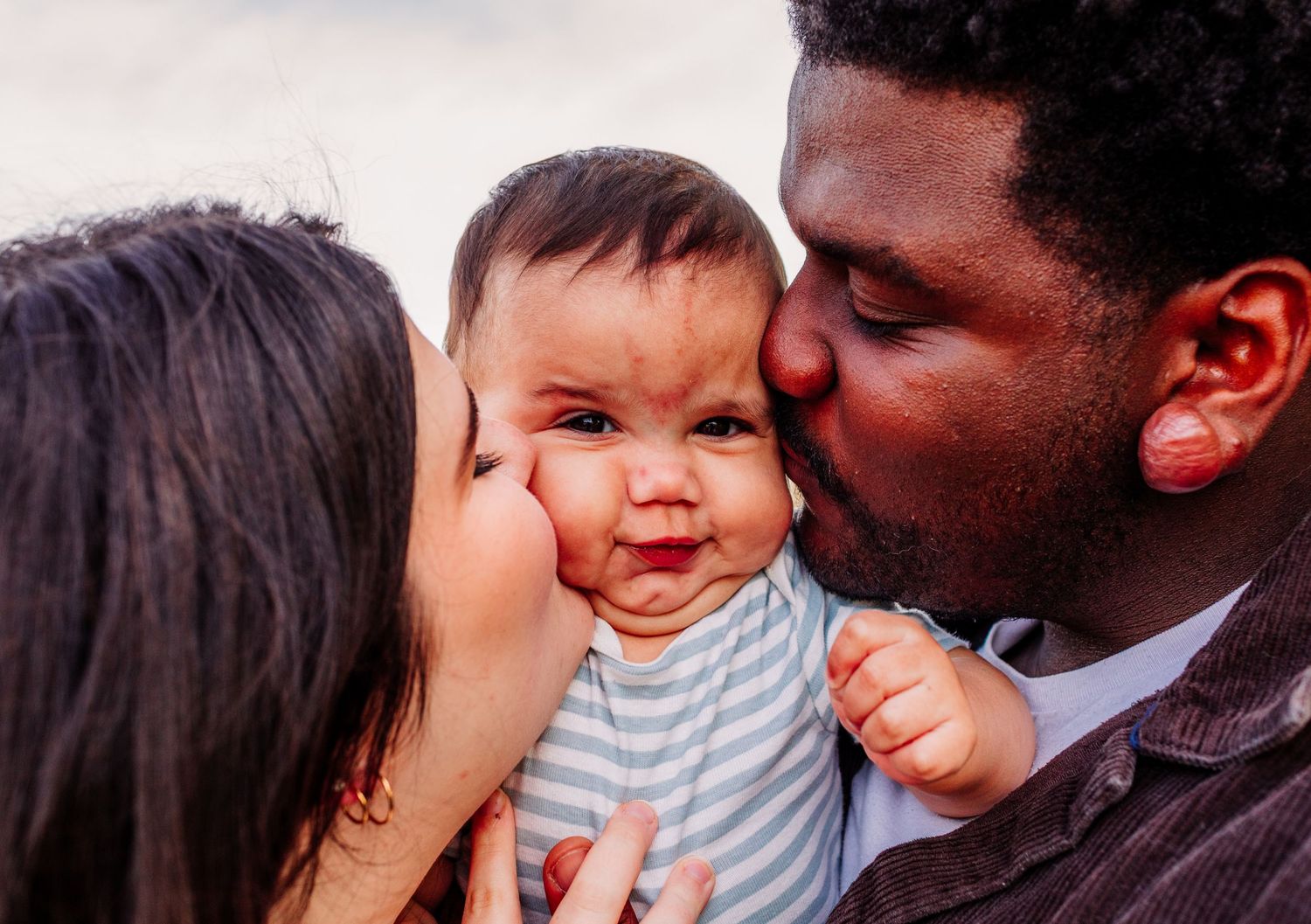Parents kissing their smiling baby on both cheeks outdoors.
