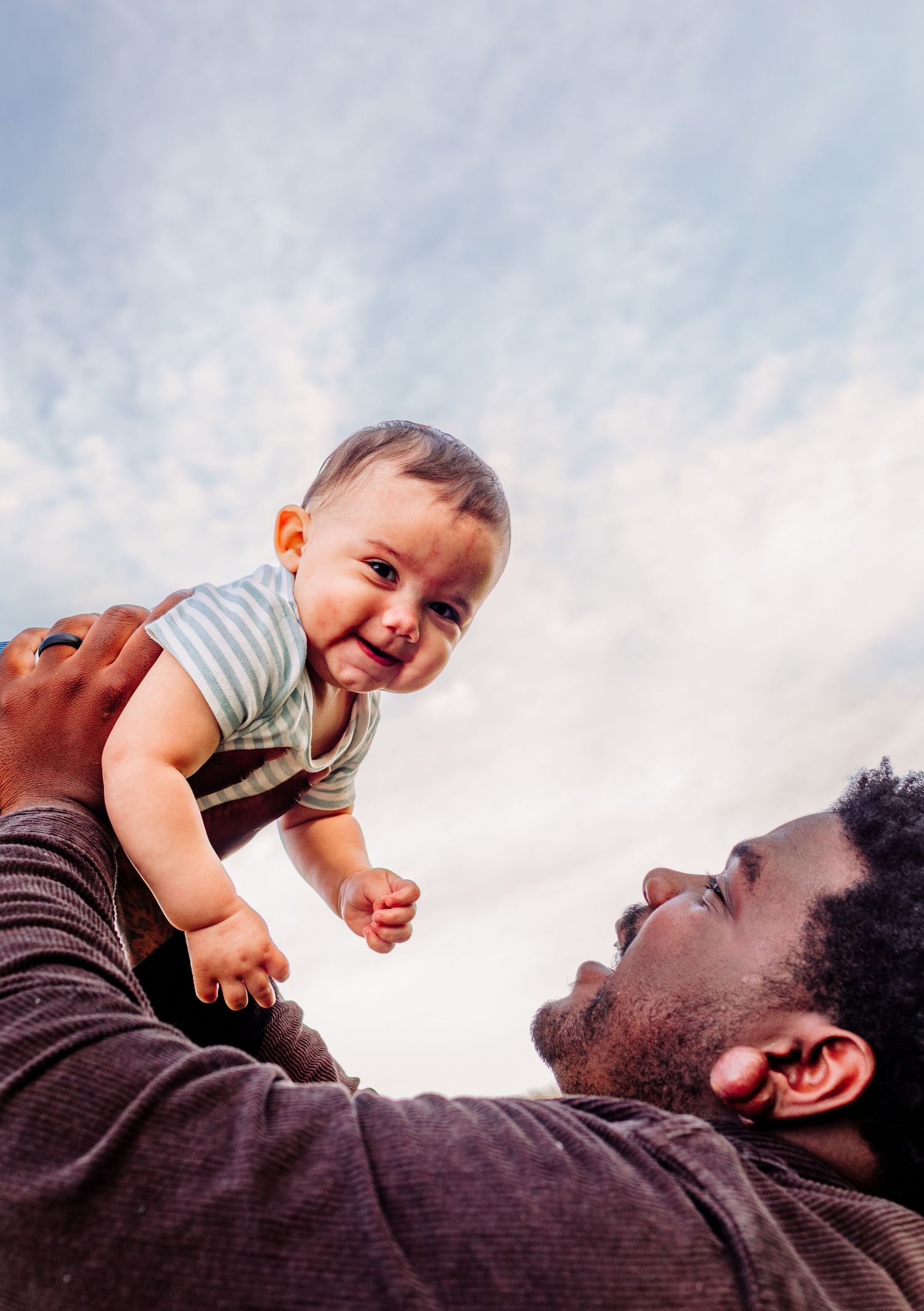Father lifting laughing baby up against cloudy blue sky during outdoor family photography session.