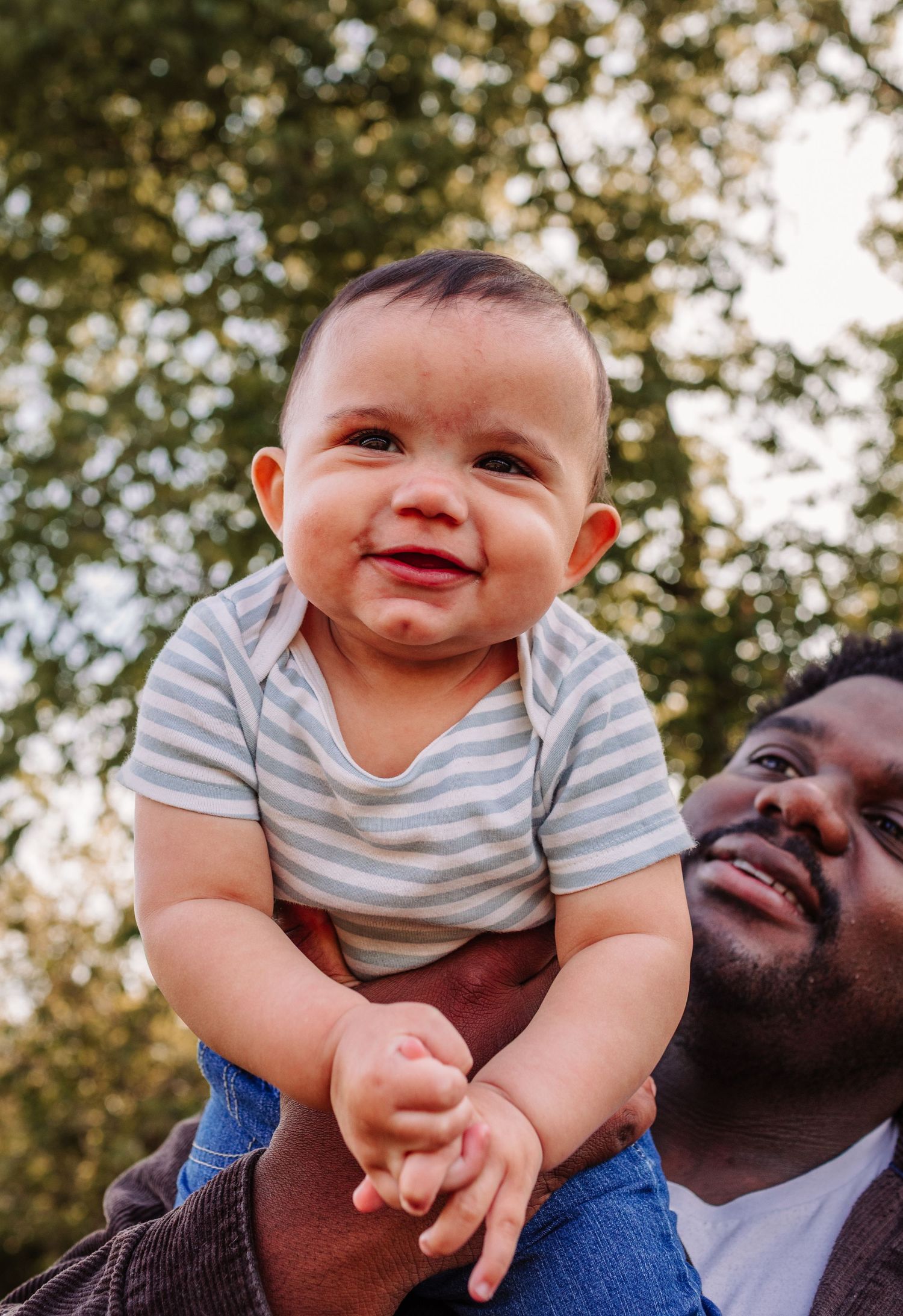Joyful baby in striped onesie lifted up by father outdoors with green trees in the background.