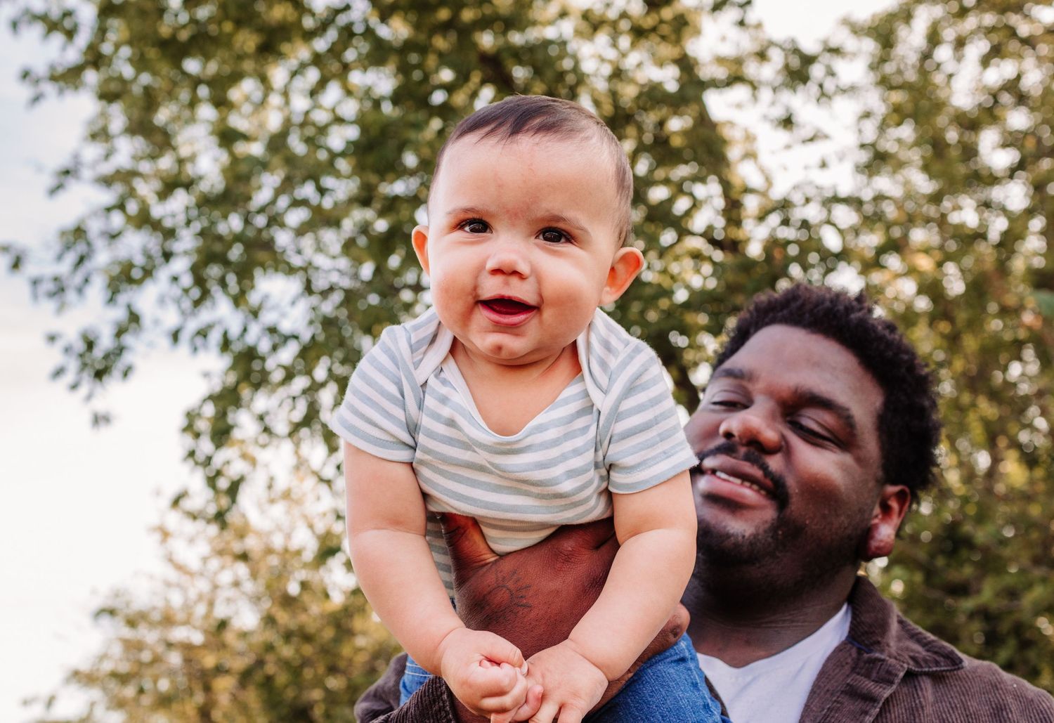 Happy father lifting smiling baby outdoors with green trees in background.