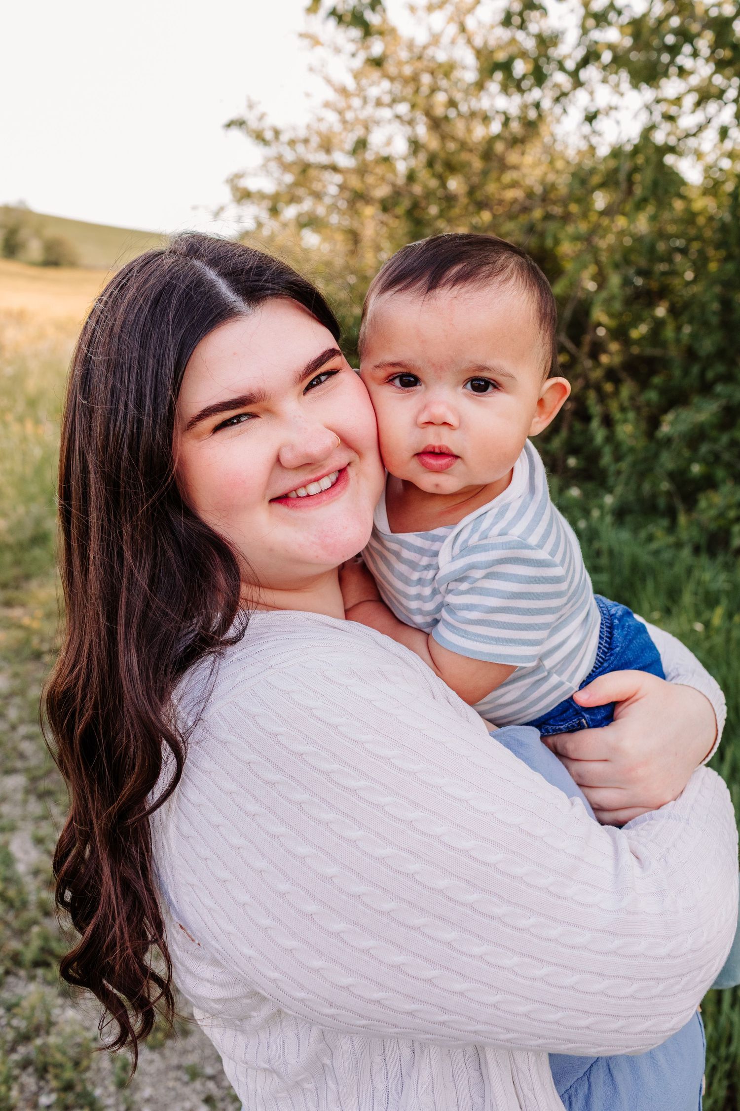 Young woman with dark hair in white sweater smiling while holding an adorable baby boy outdoors.