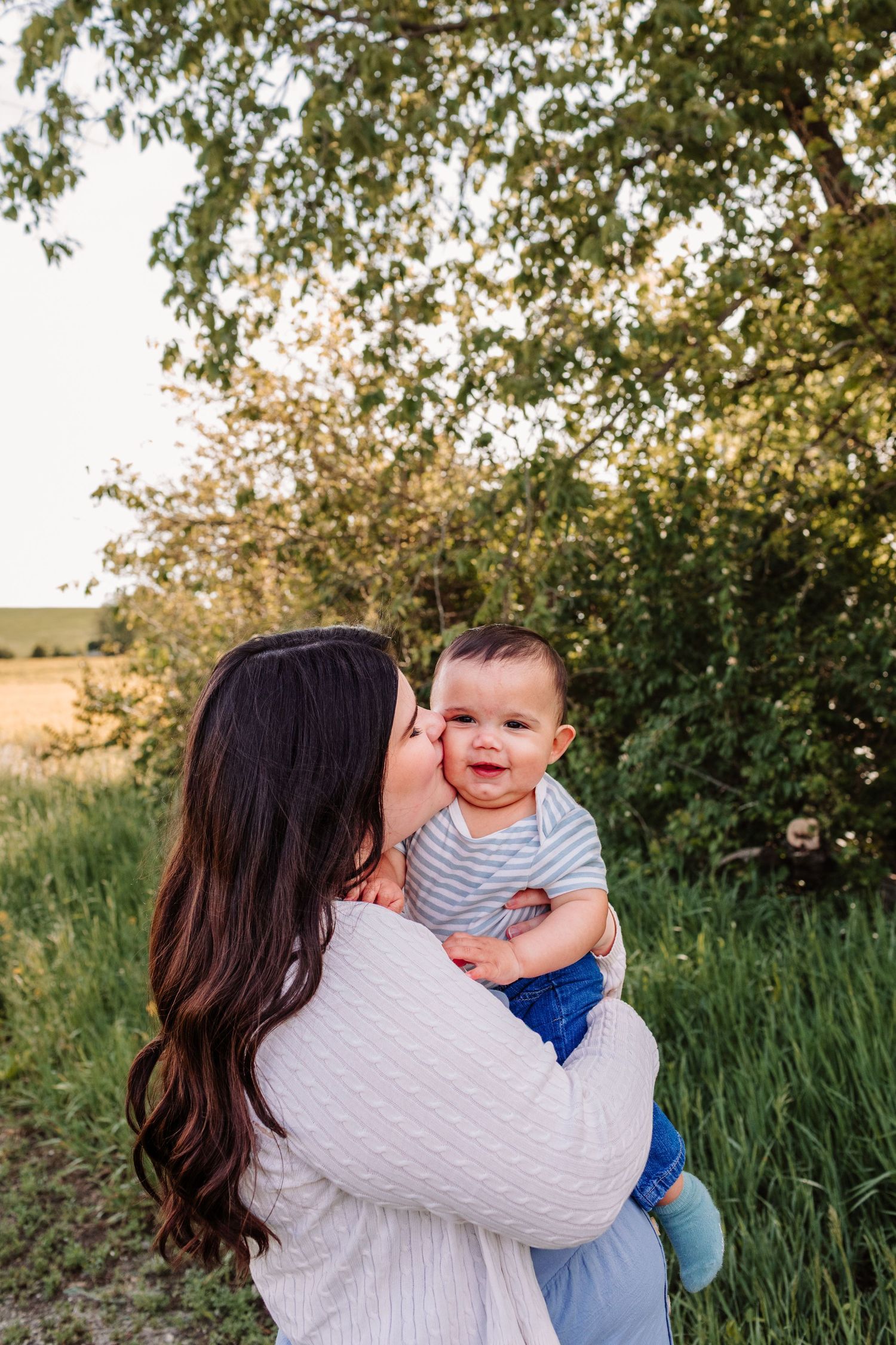Loving mother kissing smiling baby boy outdoors in lush green field during golden hour portrait.