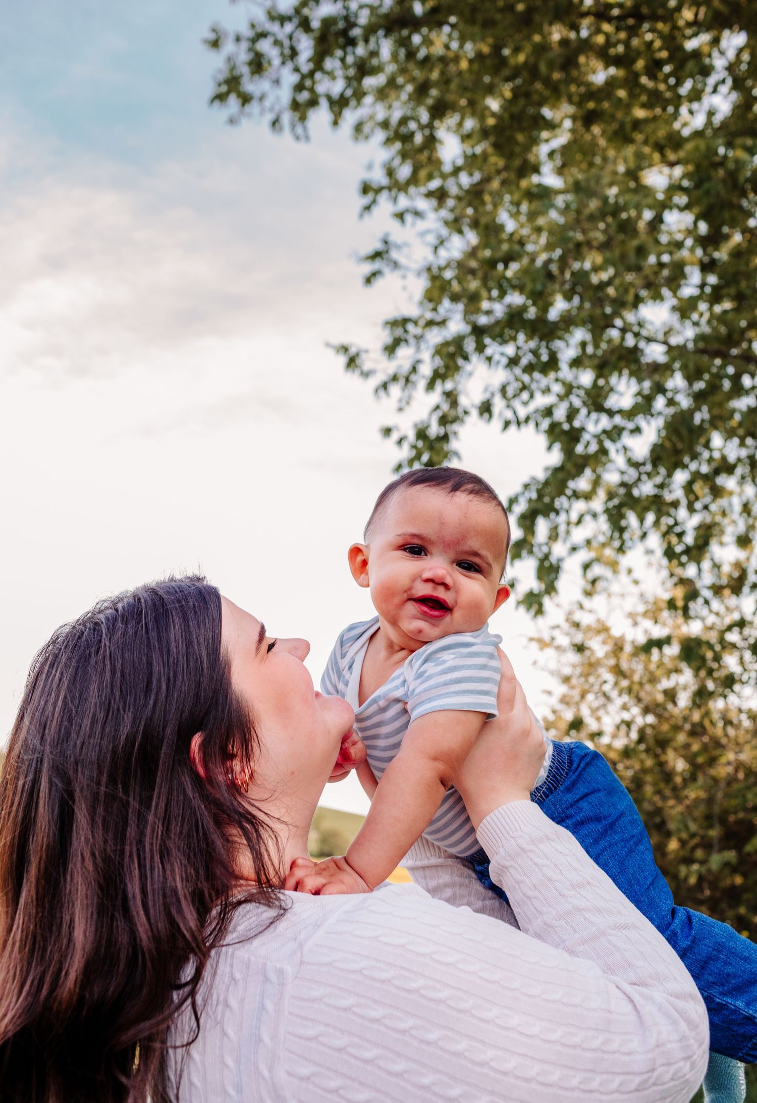 A joyful mother lifts her smiling baby outdoors with green trees and a cloudy sky in the background.