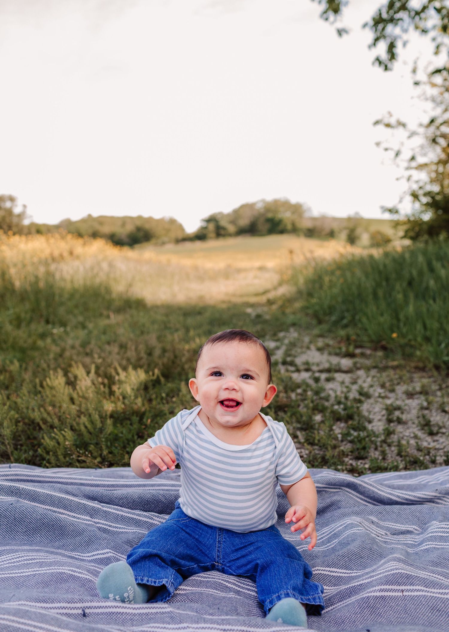 Adorable baby sitting on blanket in sunny green field smiling.