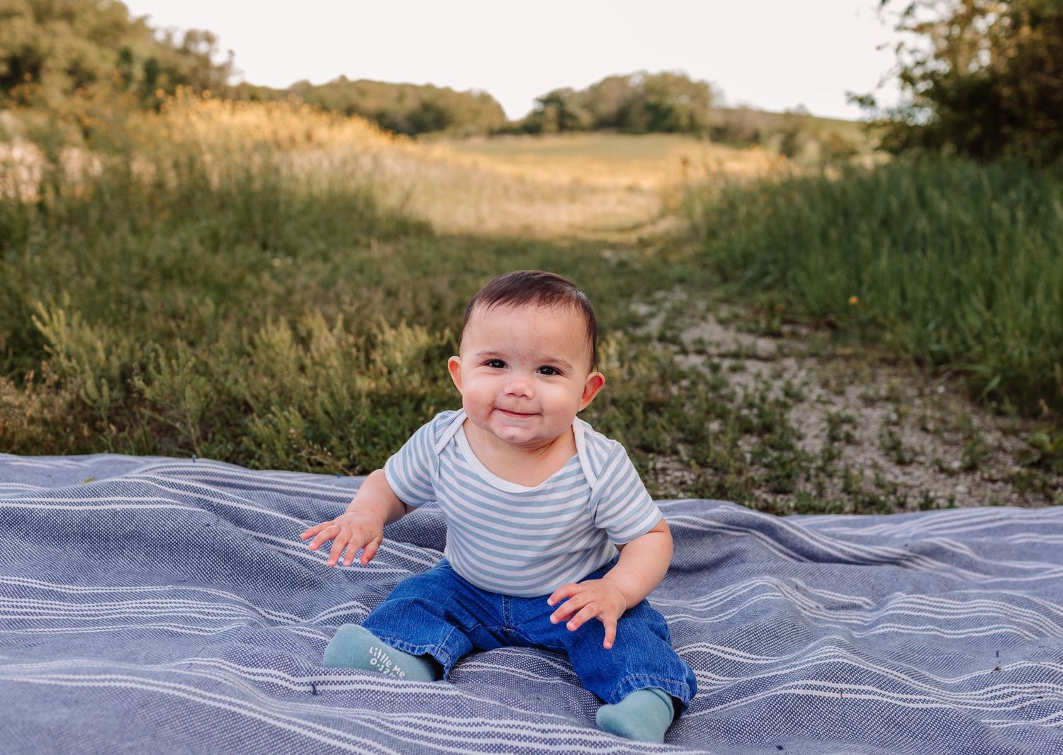 Smiling baby boy sitting on a blanket outdoors in a field wearing a striped shirt and jeans.