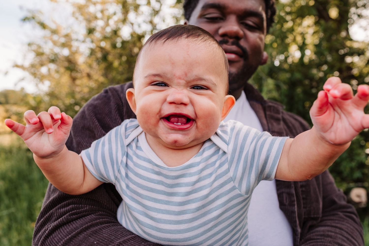 Excited baby with arms raised being held by father outdoors.