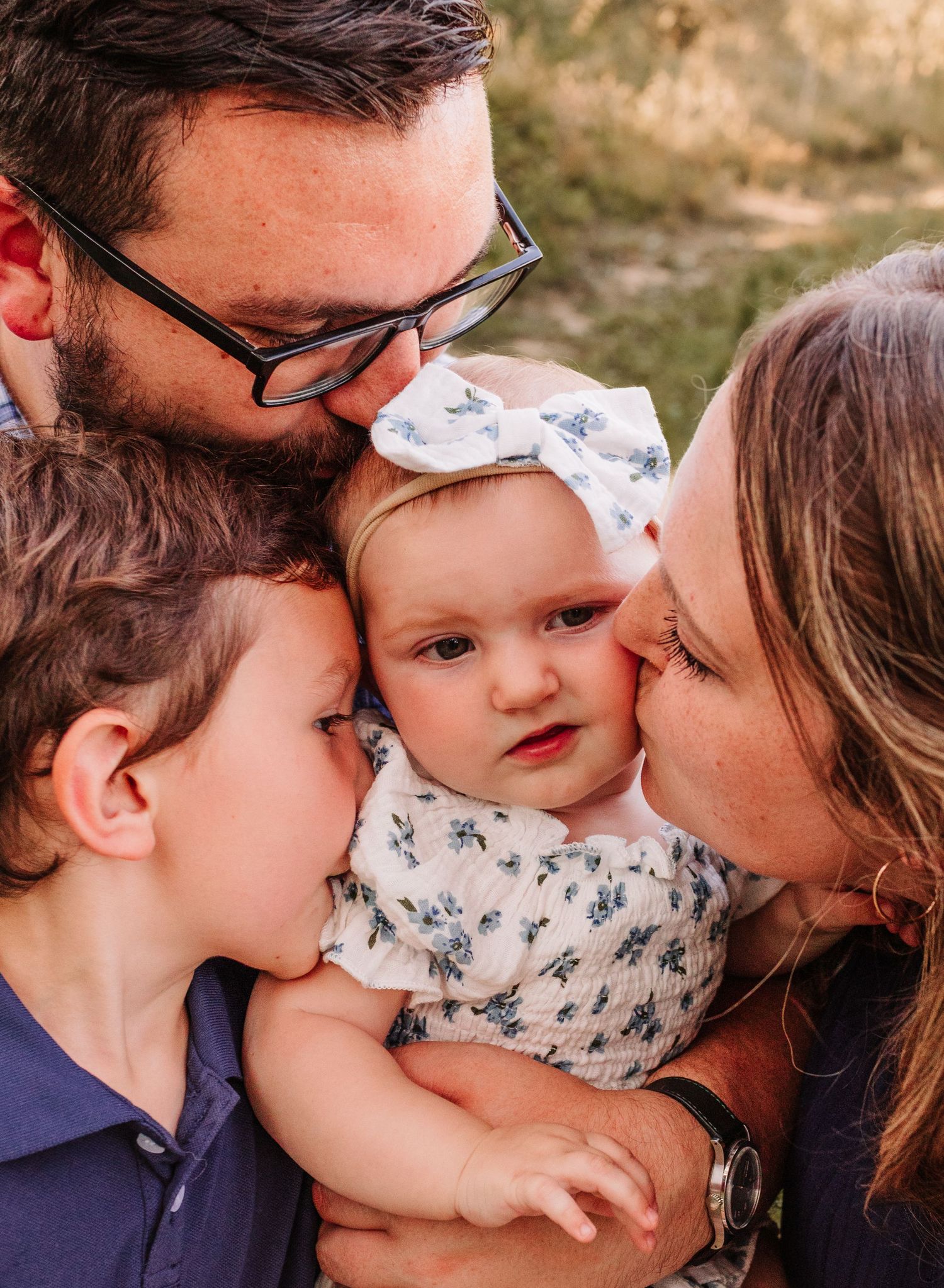 Family of four sharing a tender moment, parents and toddler kissing a baby girl wearing a white floral bow.
