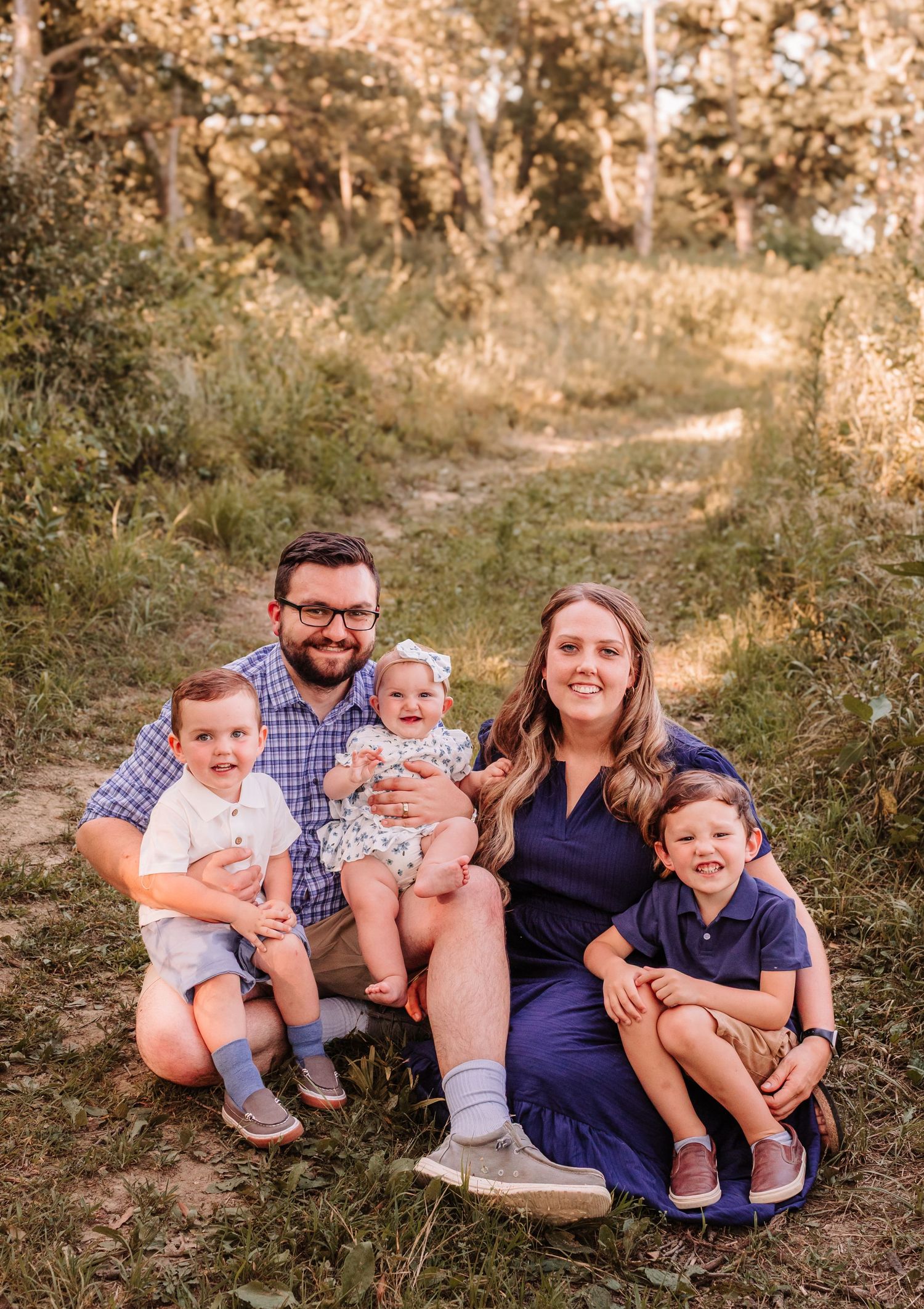 Happy family of five posing outdoors on a dirt path surrounded by lush green trees during golden hour.