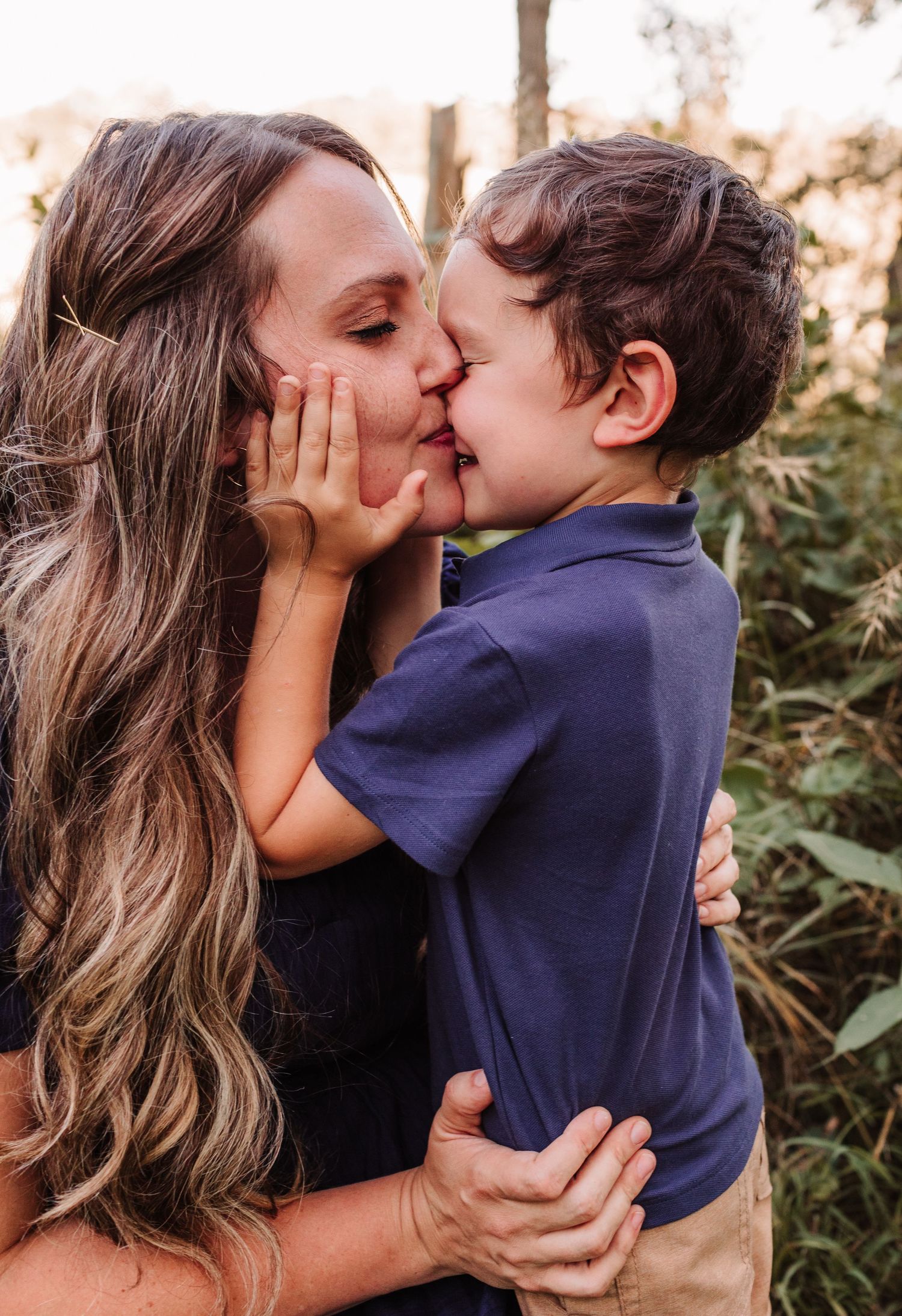 A mother and young boy share a sweet kiss outdoors, surrounded by lush greenery in a warm, golden light.