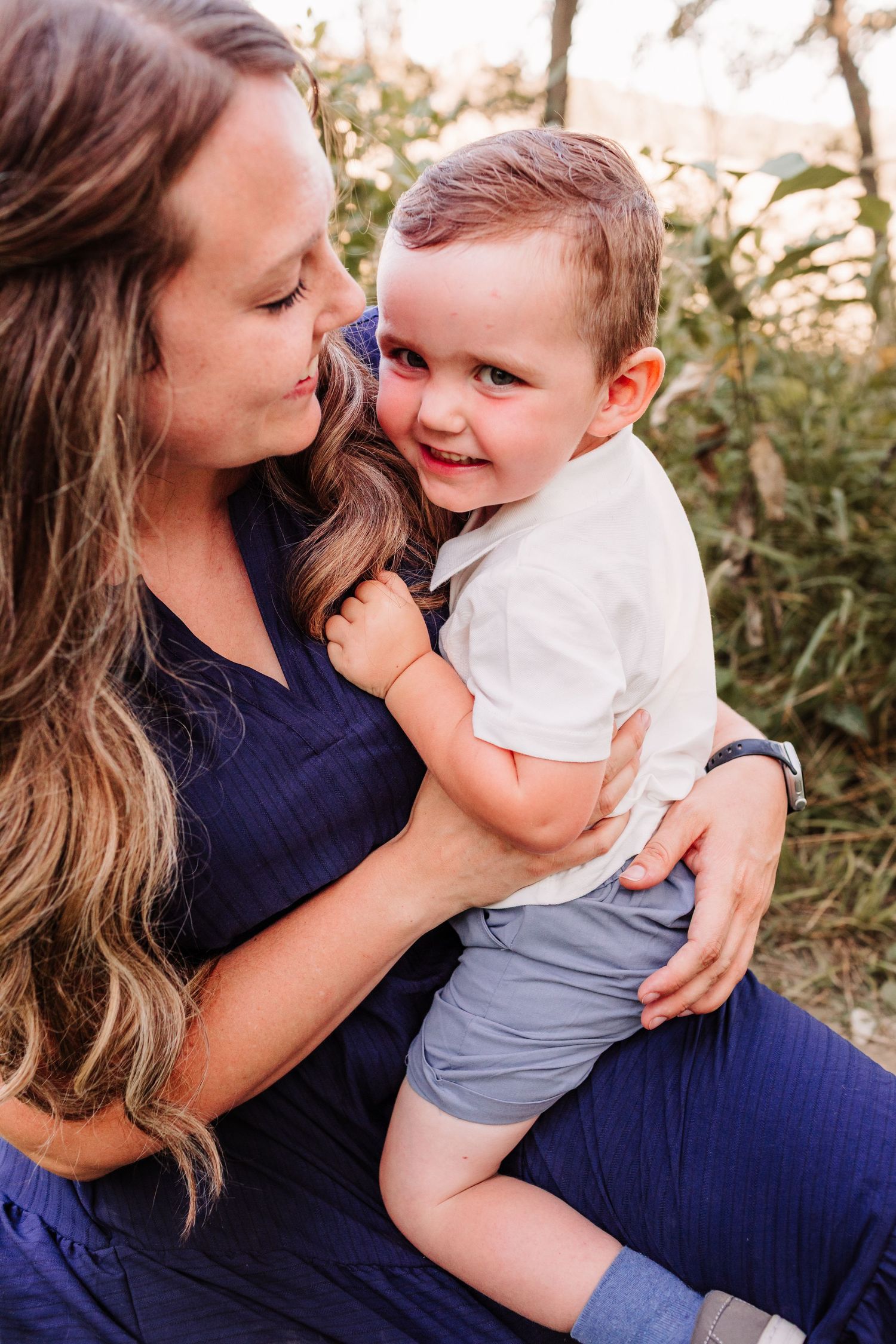 A woman in a navy dress lovingly holds a smiling toddler boy outdoors in a warm, golden-lit natural setting.