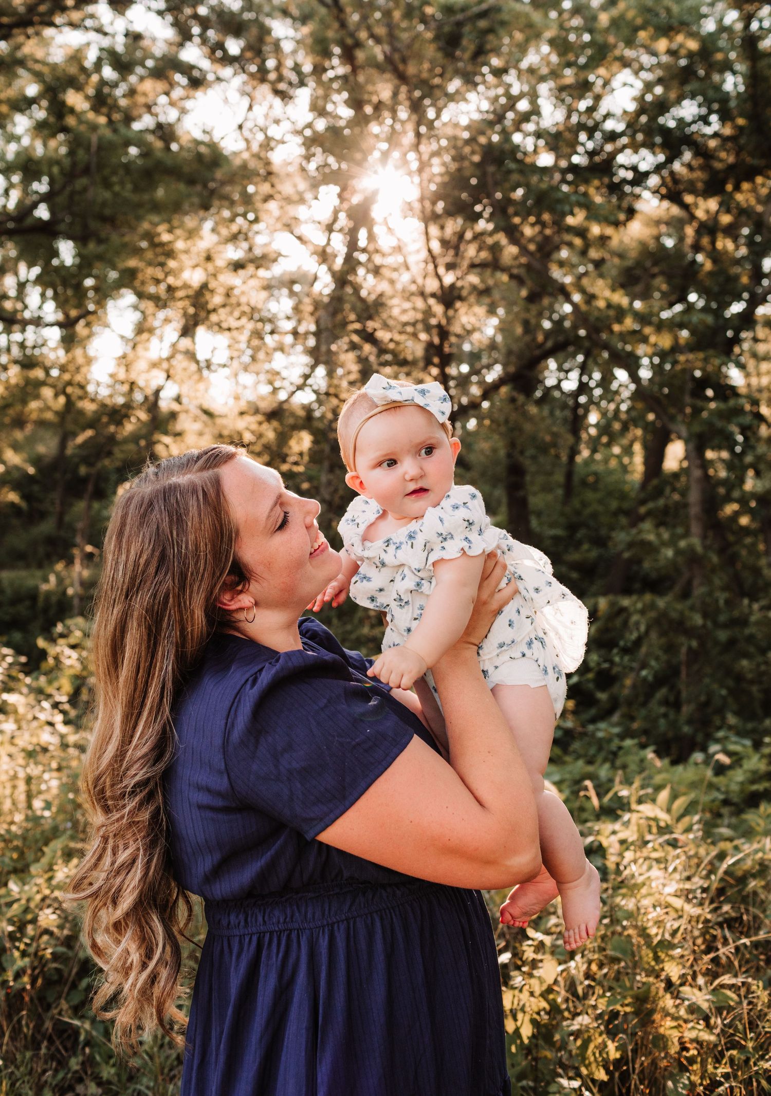 A mother in a navy dress holds her baby girl in a white floral outfit outdoors in golden hour sunlight.