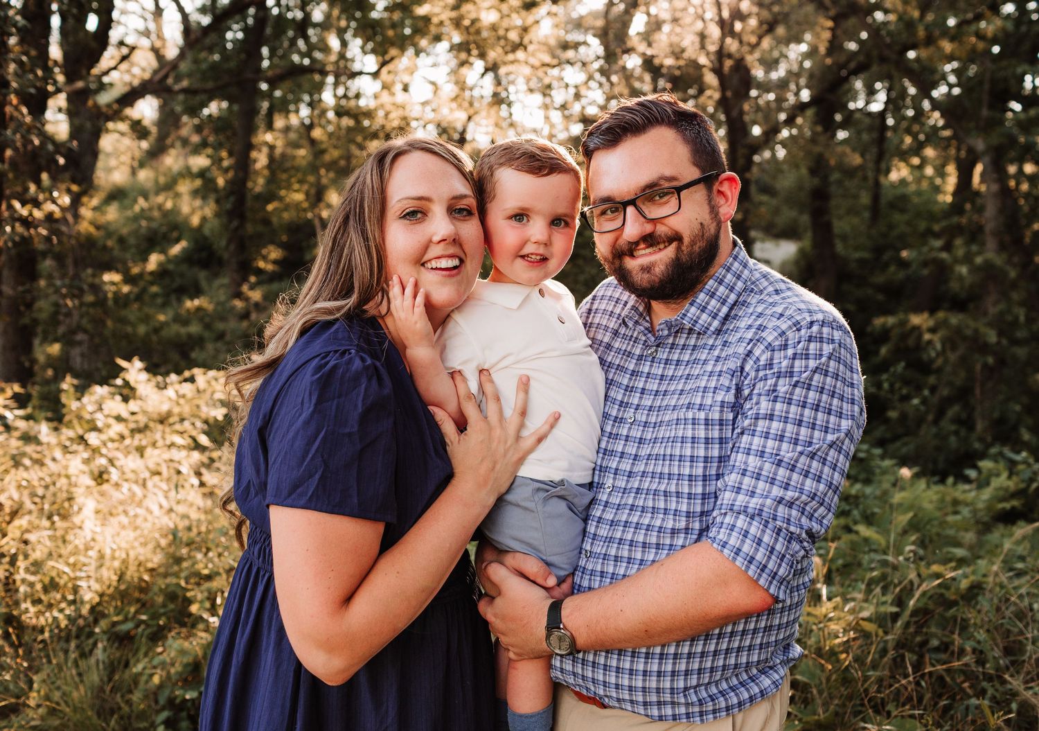 A happy young family with a toddler posing outdoors in golden sunlight surrounded by lush green trees.