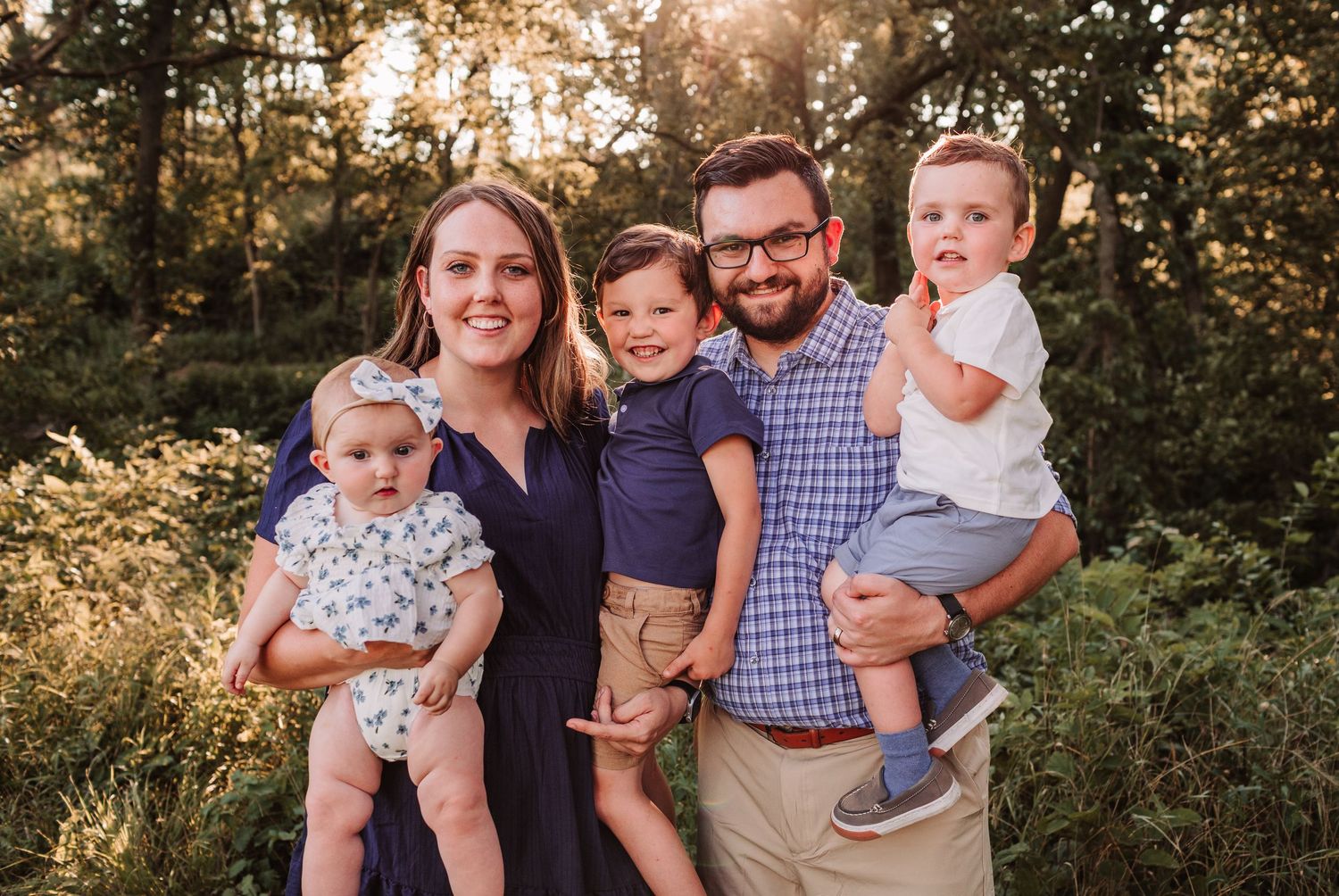 A joyful family of five with three young children smiling outdoors in warm golden hour sunlight.