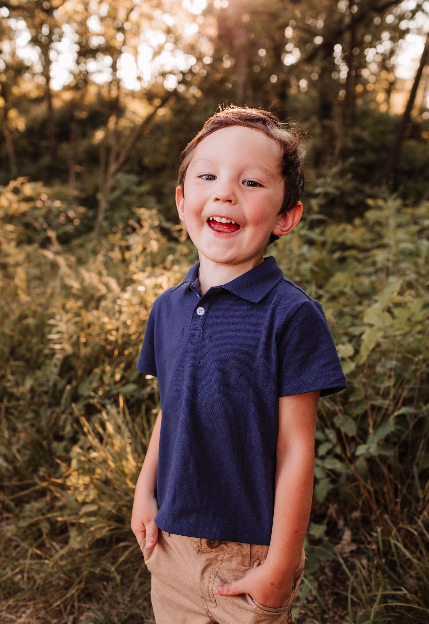 A smiling young boy in a navy polo shirt stands outdoors among lush green foliage in warm golden sunlight.