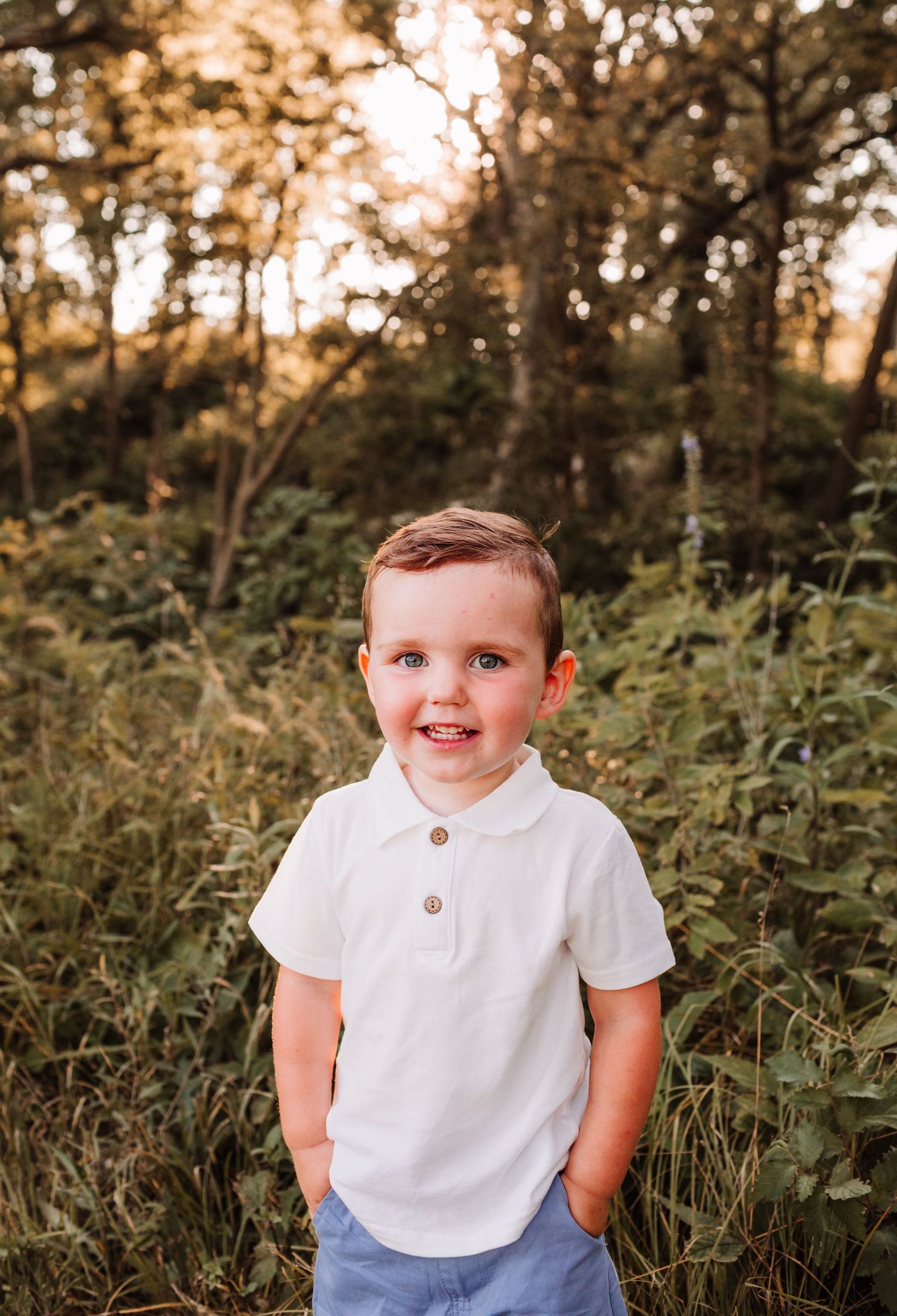 Smiling toddler boy in white polo shirt standing in lush green field with golden sunlight filtering through trees.