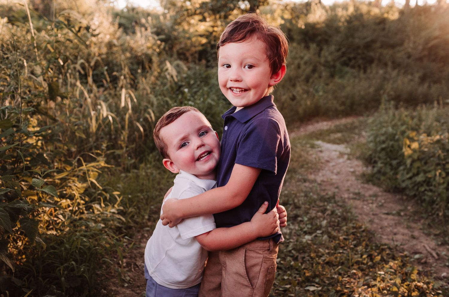 Two young brothers hugging and smiling on a dirt path surrounded by lush greenery in warm golden hour light.
