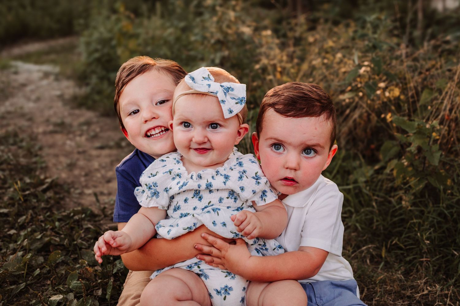 Three young siblings pose outdoors, two boys flanking a baby girl in a floral dress with a bow headband.