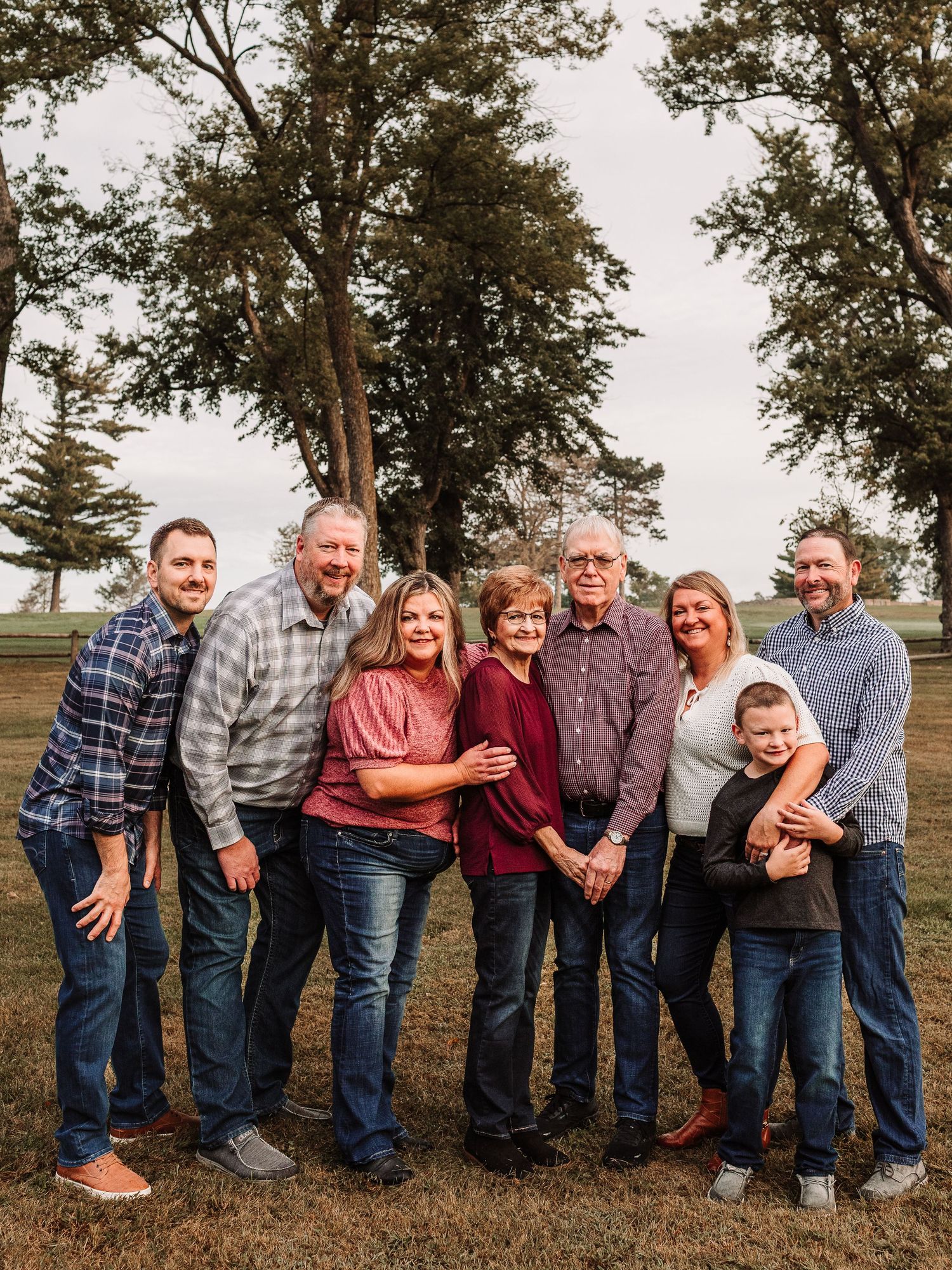 Multi-generational family of eight posing together outdoors in a park with tall trees in the background.