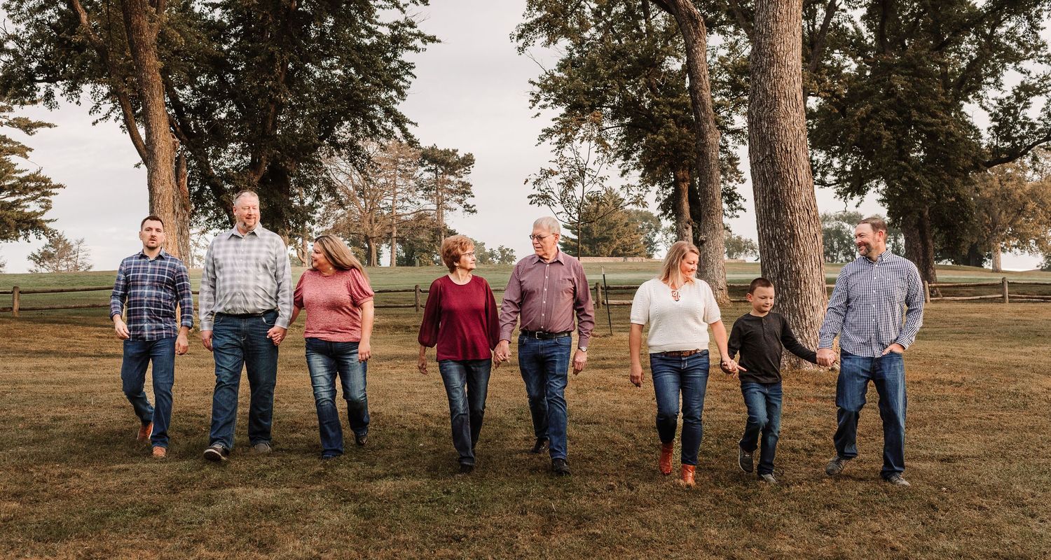 Extended family of eight walking together hand-in-hand across a green lawn with large trees behind them.