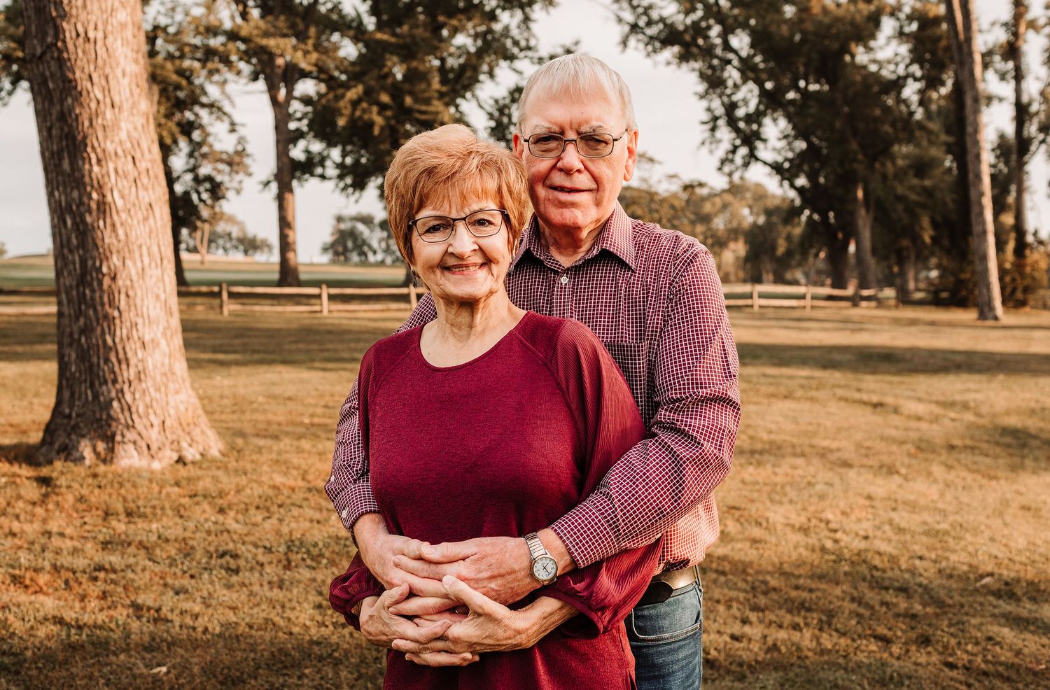 Smiling older couple embracing outdoors, woman in burgundy top and man in plaid shirt, standing in a park.