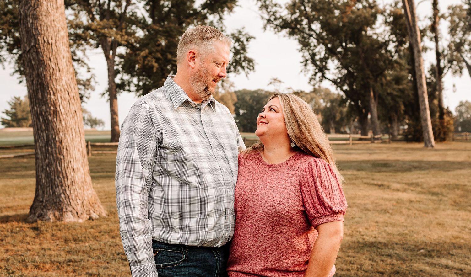 Middle-aged couple smiling at each other outdoors in a park, man in plaid shirt and woman in pink top.