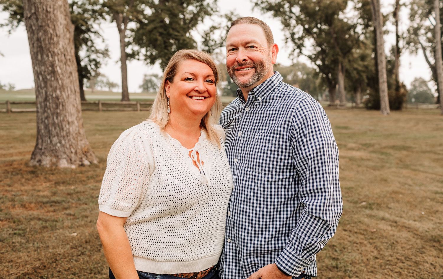 Smiling couple posing together outdoors in a park, woman in white knit top and man in blue gingham shirt.