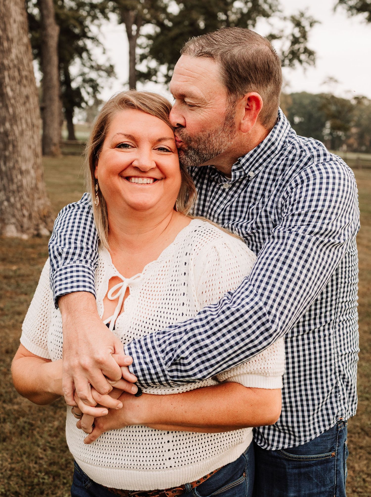 Man in blue gingham shirt kissing smiling woman on the cheek while embracing her from behind outdoors.