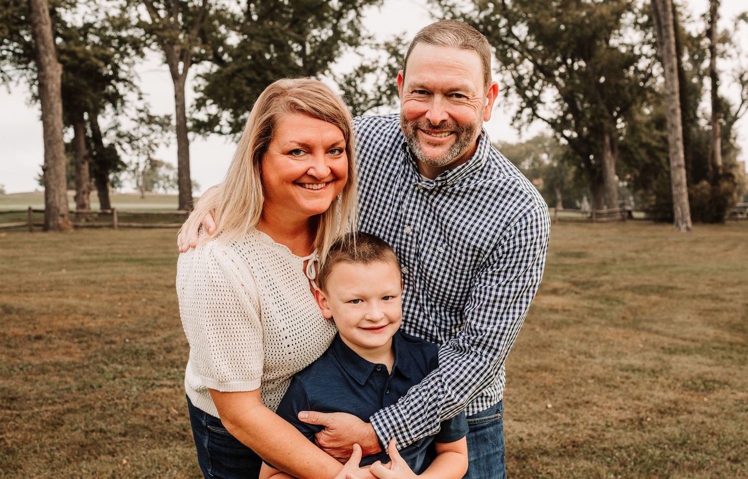 Smiling family of three with young boy posing together outdoors in a grassy park with trees in background.