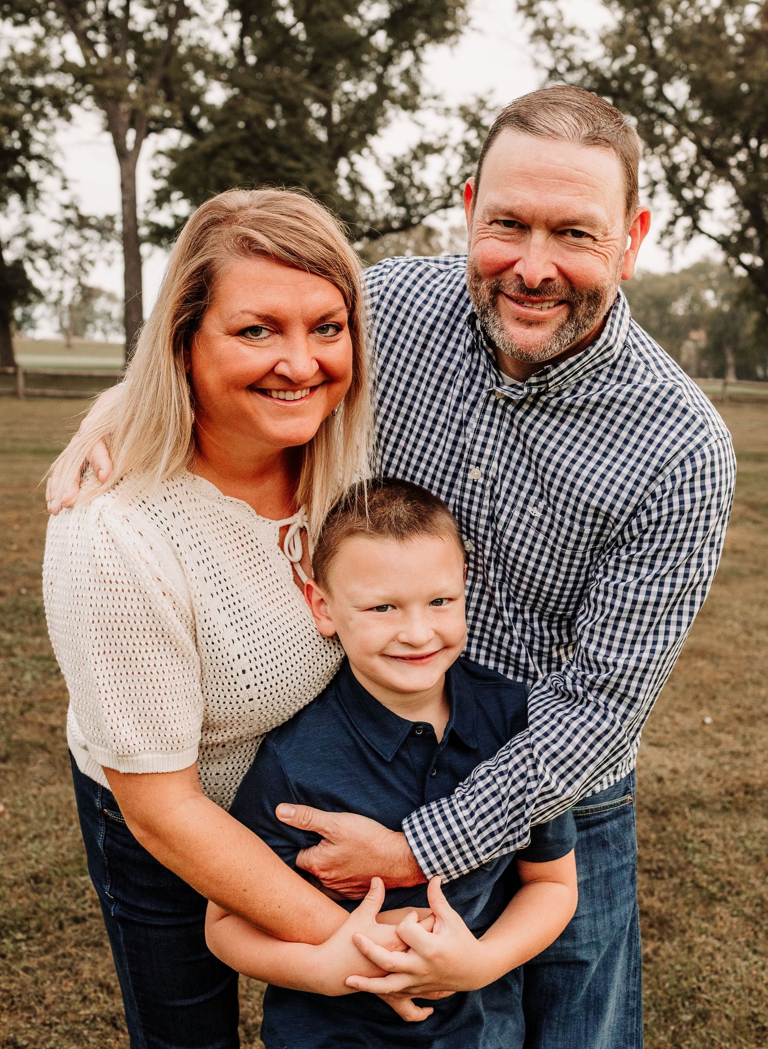 Happy family portrait of parents and young son smiling together outdoors in a park with green trees behind.