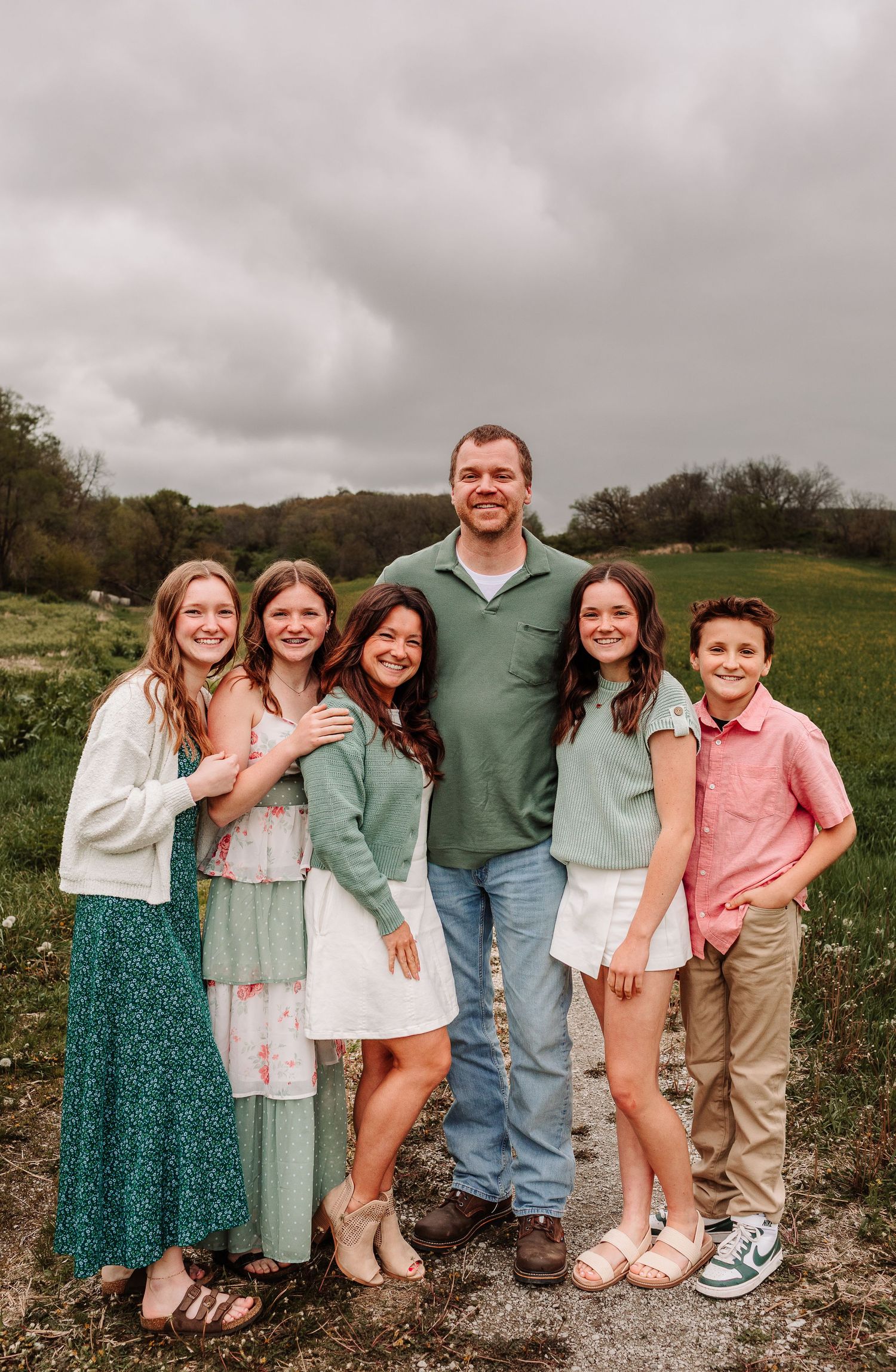 Family of six posing outdoors in a green field wearing coordinated pastel outfits under a cloudy sky.