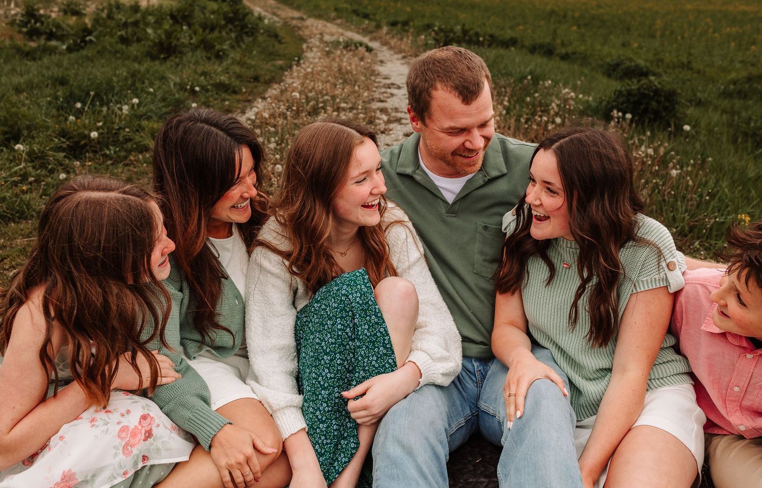 A joyful family of six laughing together outdoors in a green meadow, wearing coordinated sage green and white outfits.