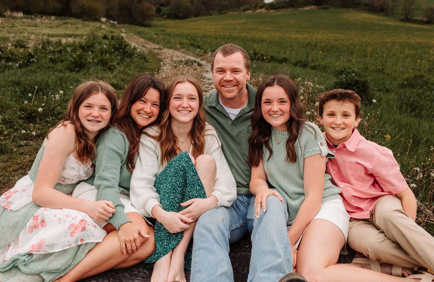 Happy family of six posing together in a lush green field, wearing coordinated sage green, white, and pink outfits.