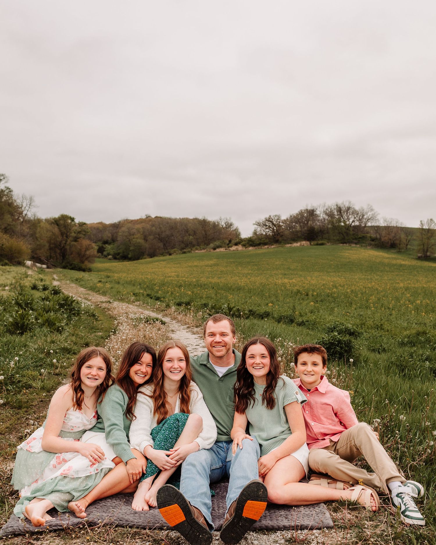 Family of six smiling together on a blanket in a green field on a cloudy day.