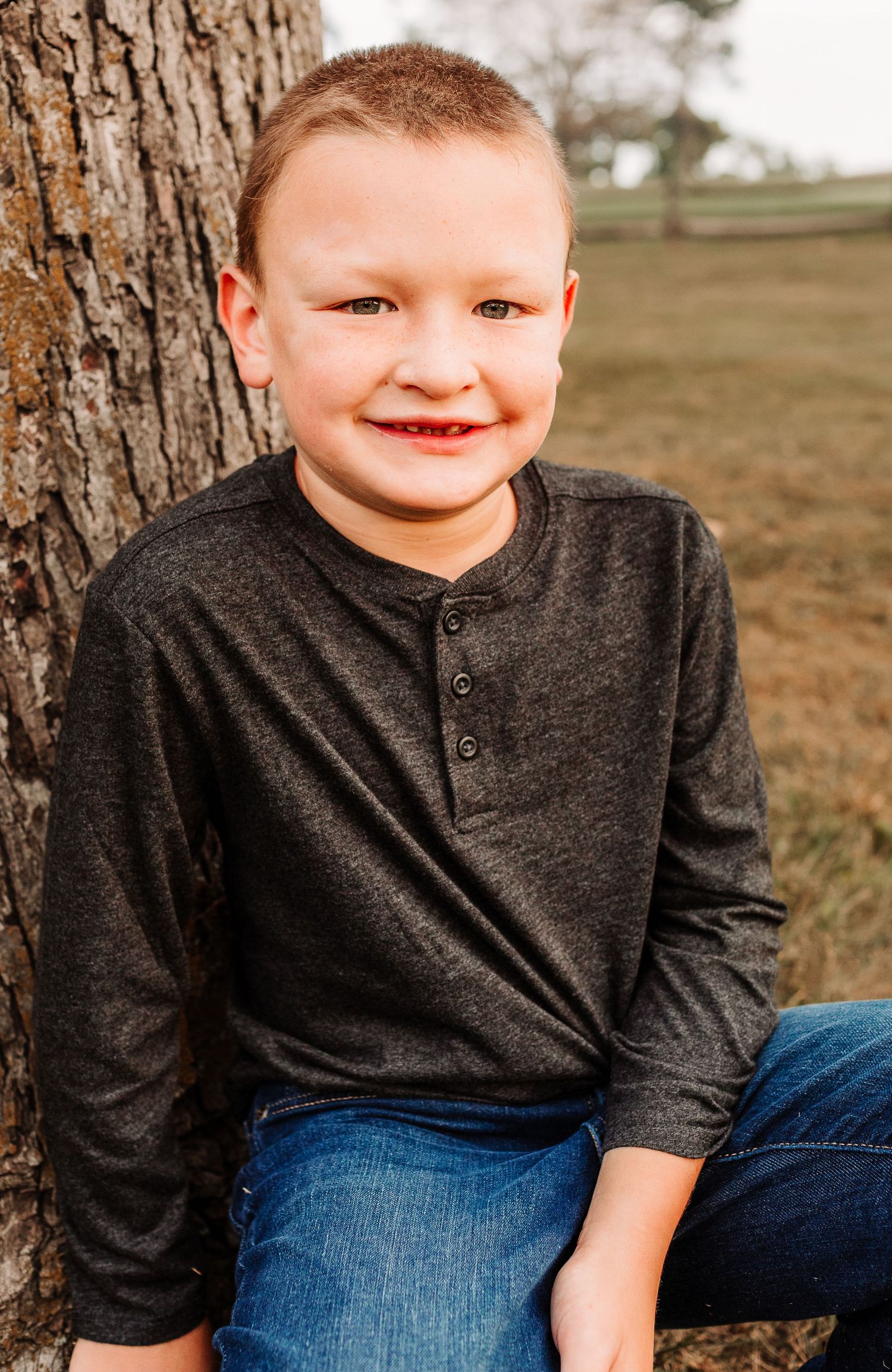 Smiling young boy in dark henley shirt leaning against tree outdoors.