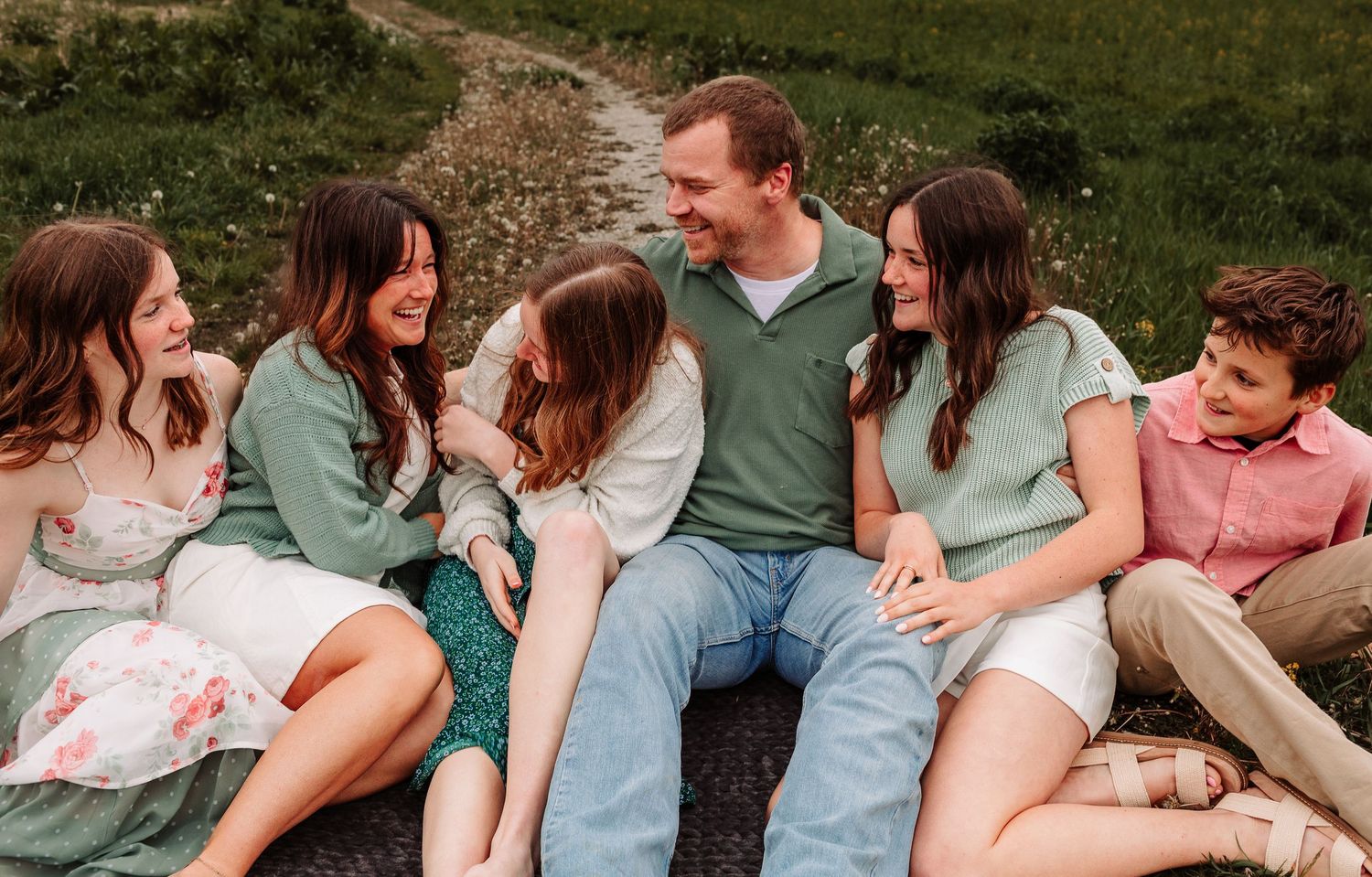 Happy family of five sitting together outdoors on a grassy hillside, smiling and laughing in coordinated green outfits.