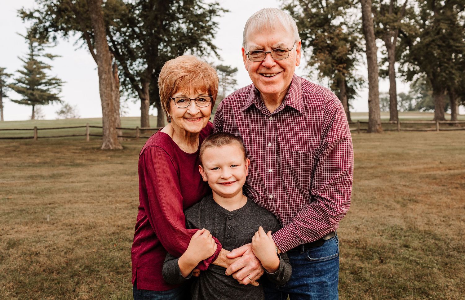 Grandparents hugging young grandson outdoors in park setting.