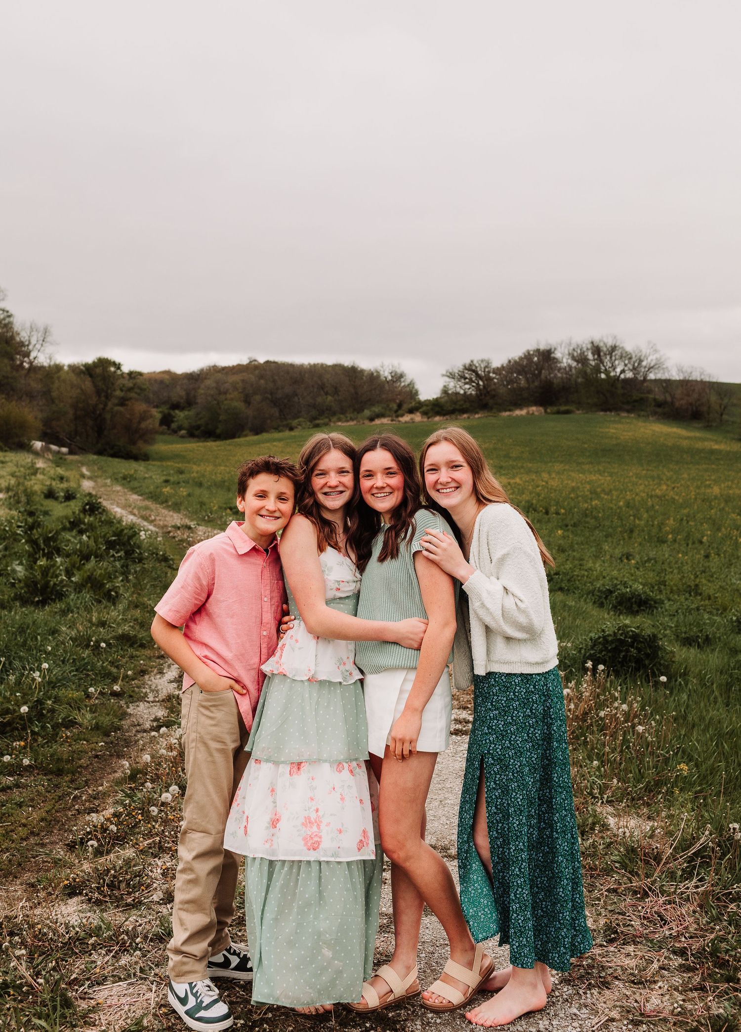 Four smiling children pose together on a dirt path surrounded by green fields on a cloudy day.