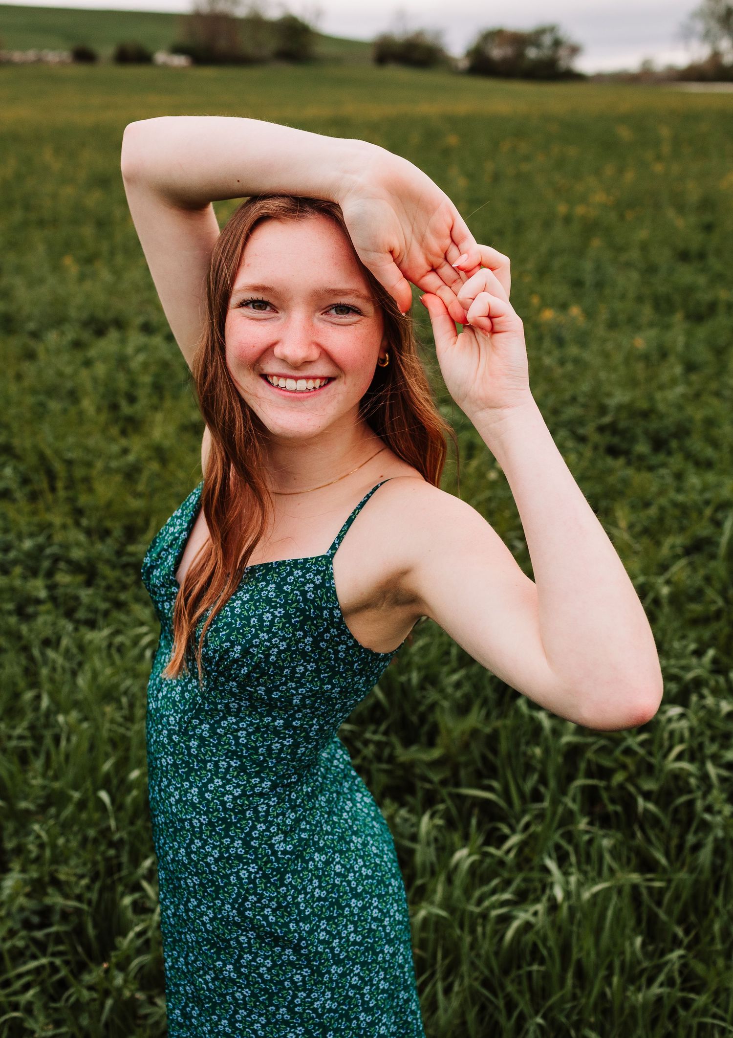 Young woman with red hair smiling in a green floral sundress, posing in a lush green field.