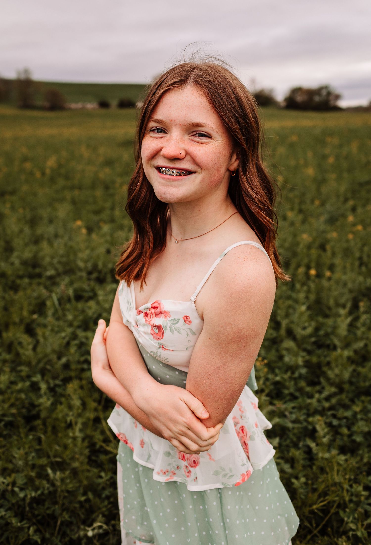 Smiling teenage girl with braces in floral dress standing in green field on cloudy day.