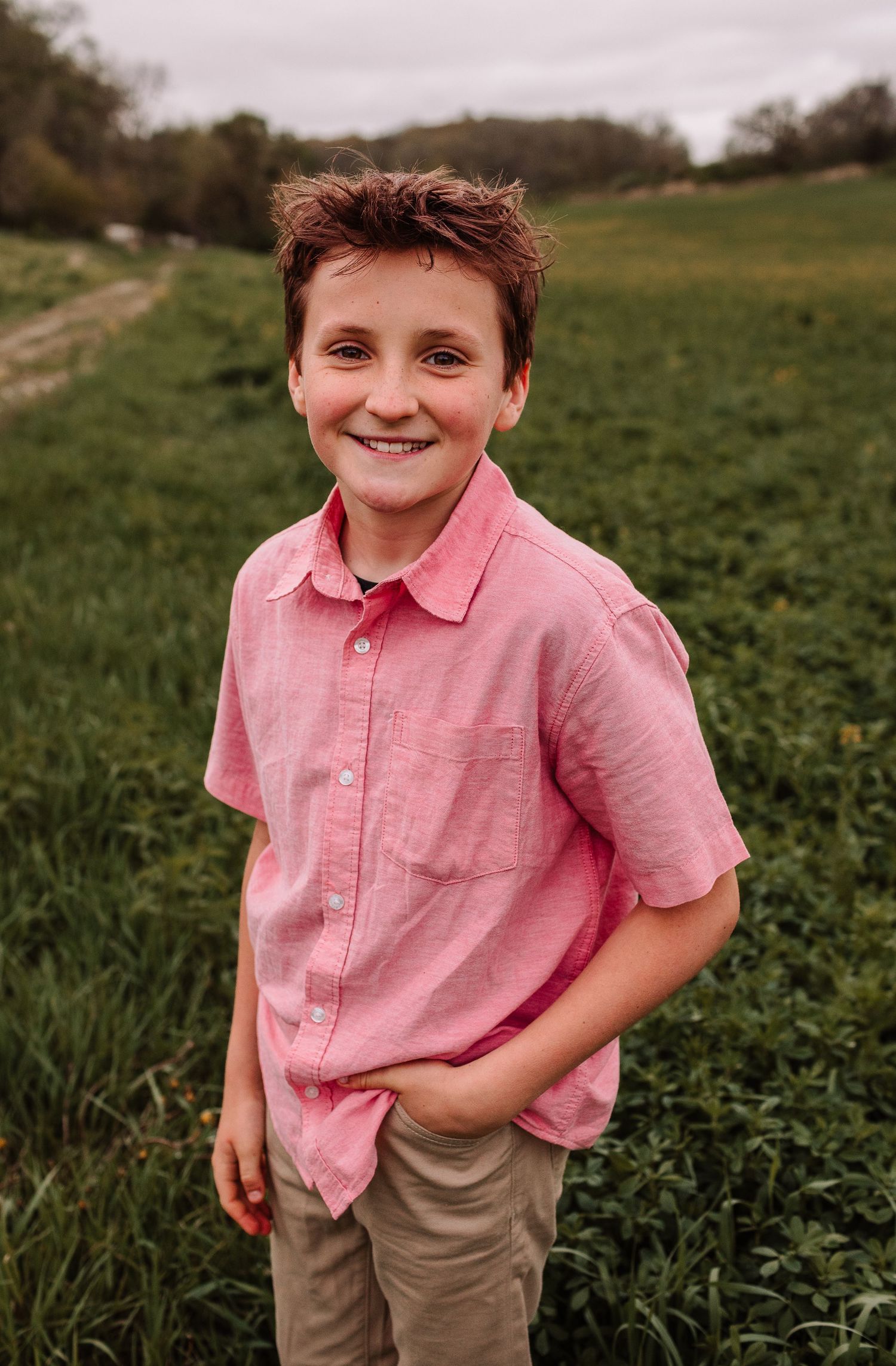Smiling boy in pink short-sleeve shirt standing in a green field, hands in pockets, posing for a portrait photo.