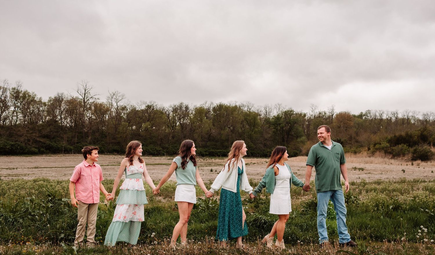 Family of six holding hands in a line walking through a green meadow.