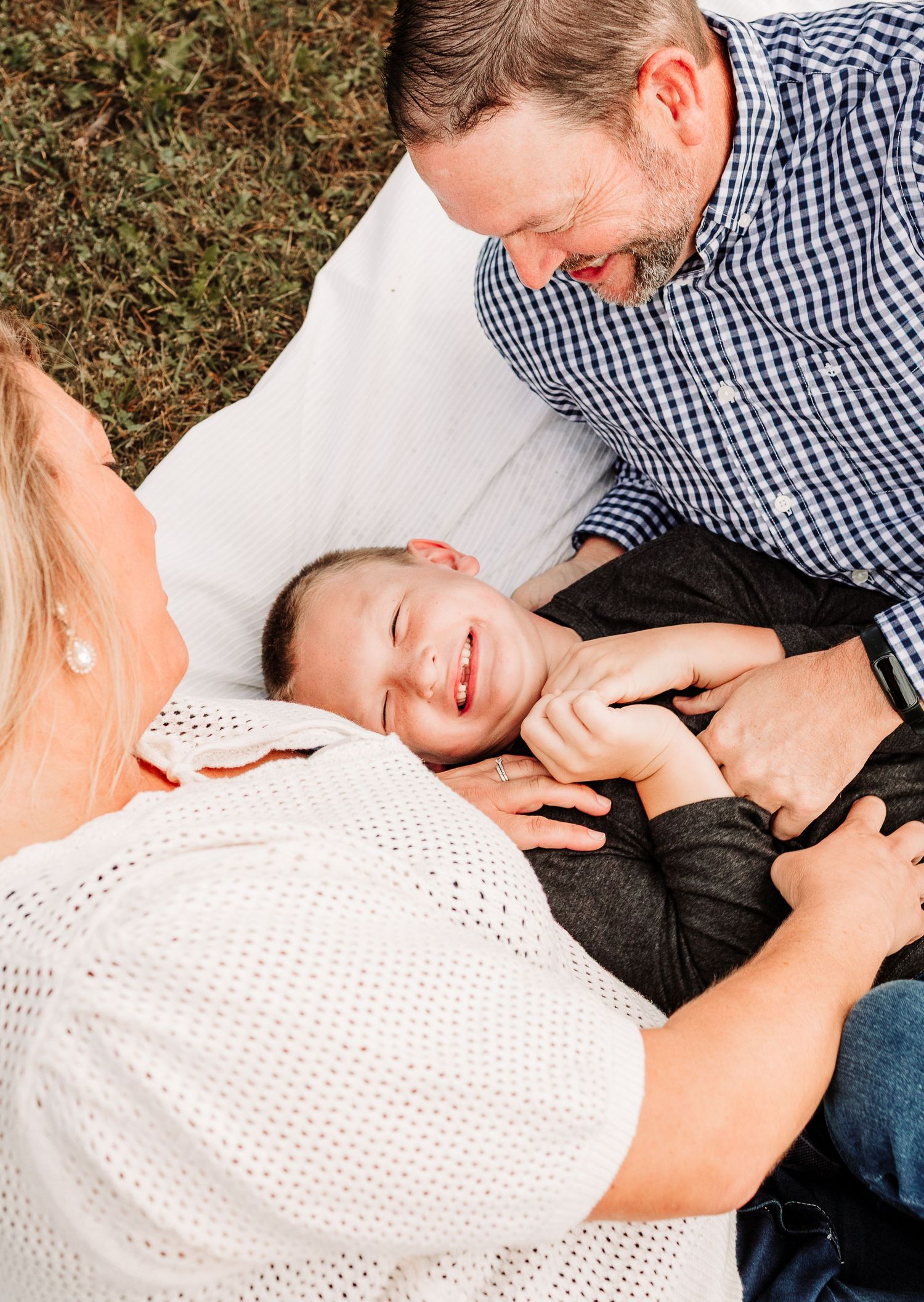 Family laughing together on blanket outdoors, parents playing with young boy.