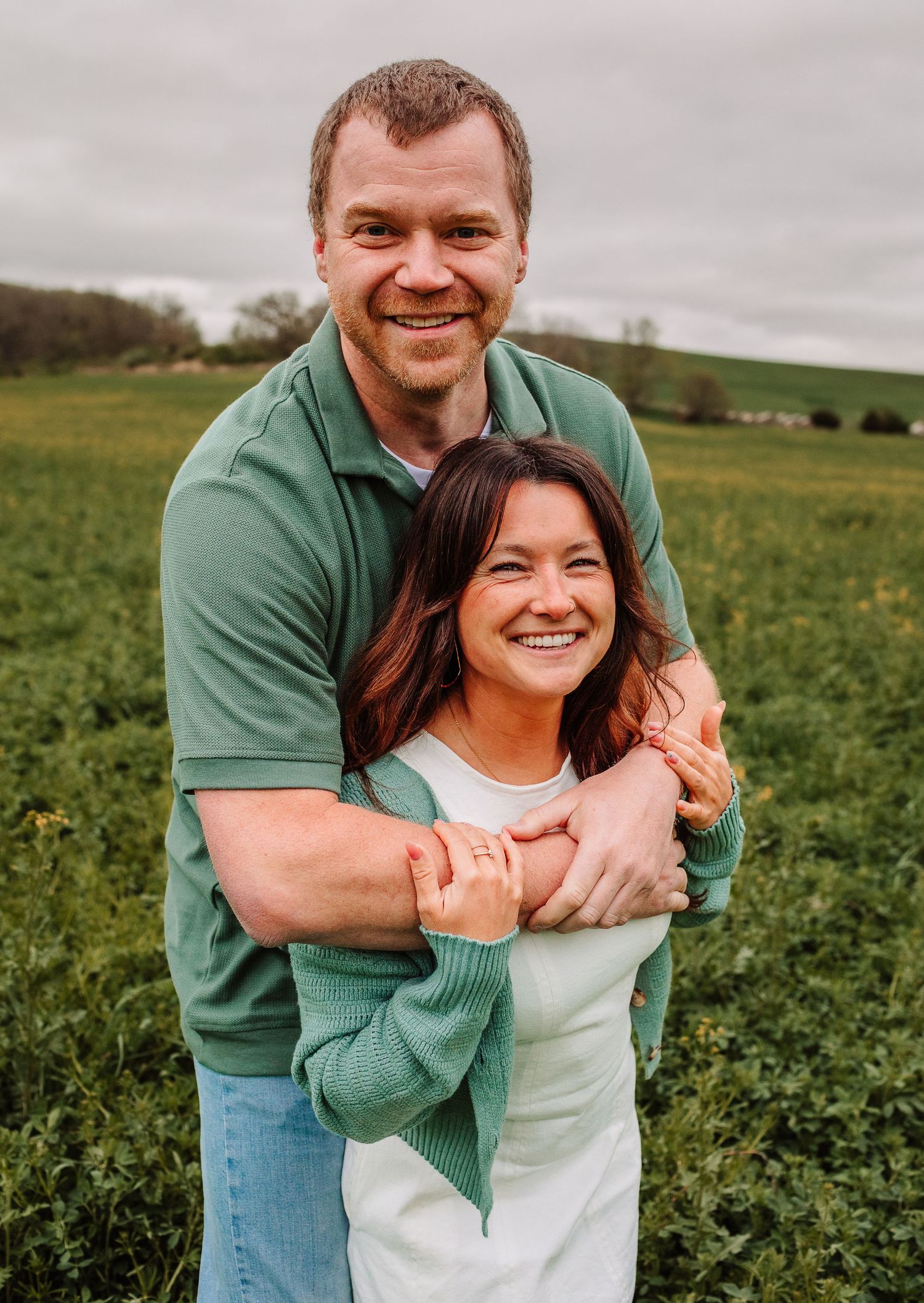 Smiling couple embracing in a green field, man in green polo hugging woman from behind.
