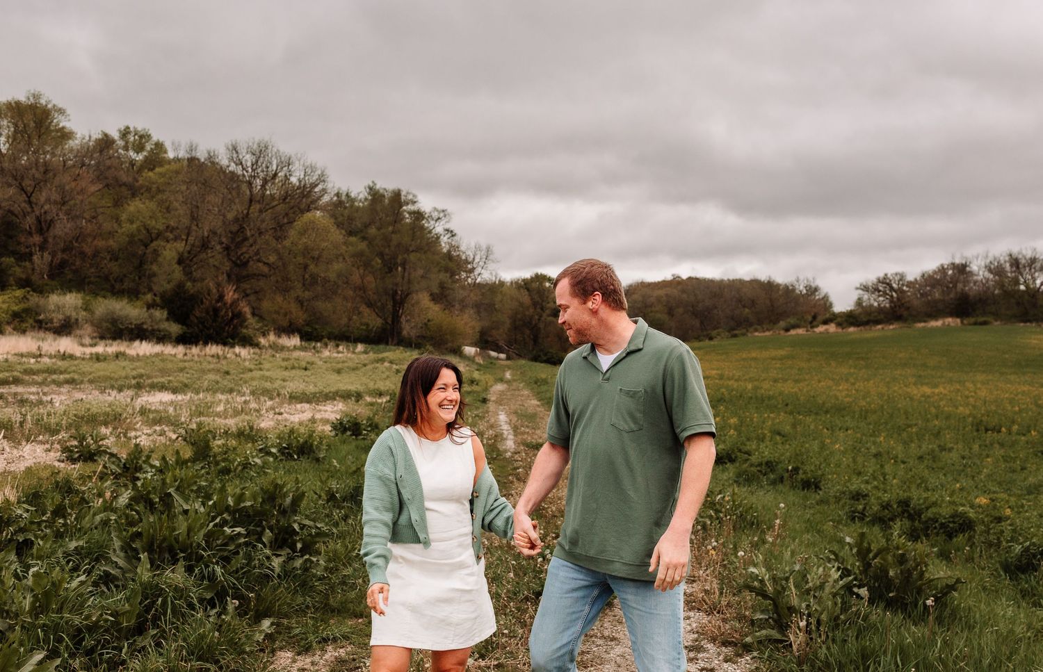 Couple holding hands smiling at each other in a green field with trees and cloudy sky backdrop.