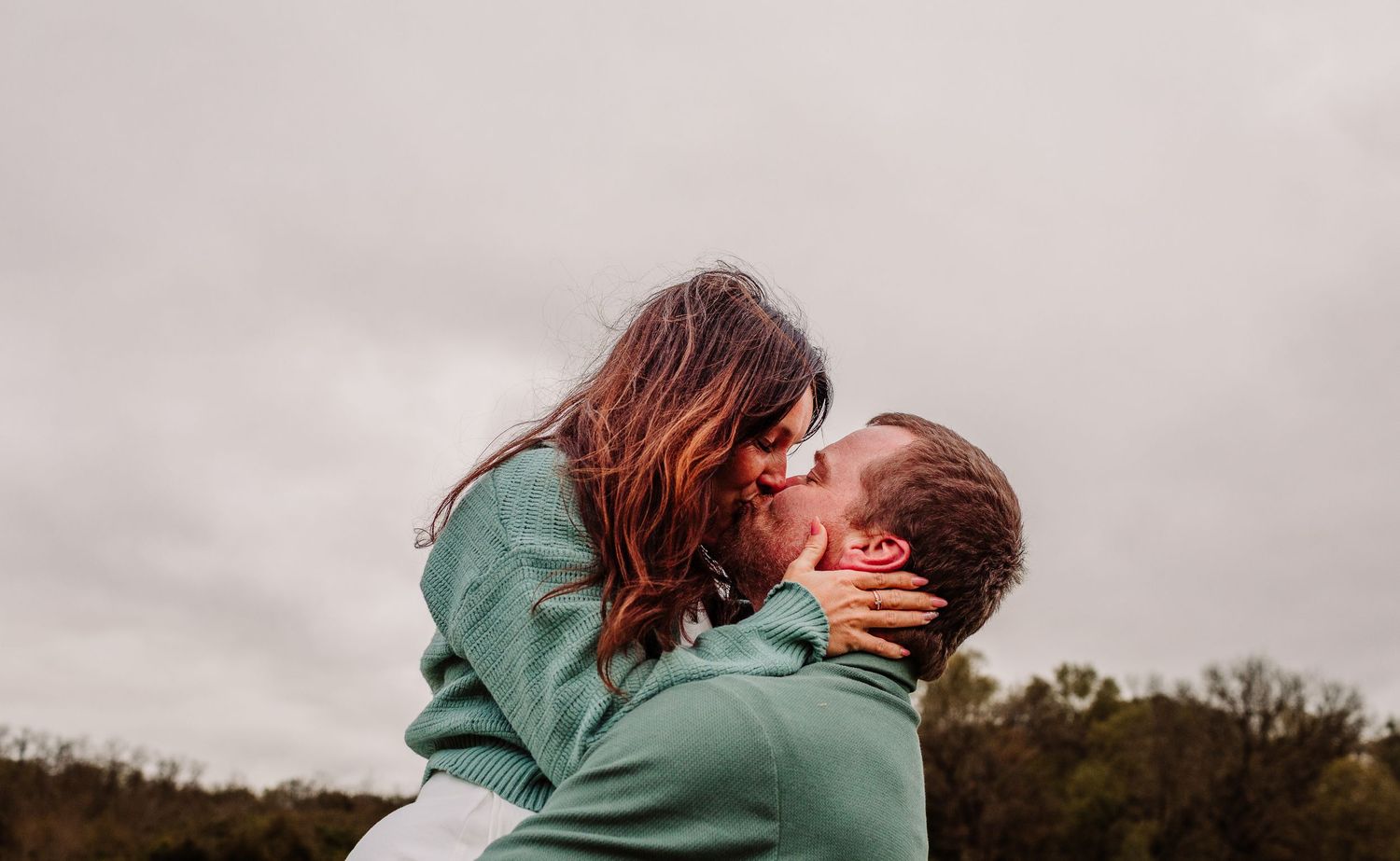 A couple sharing a romantic kiss outdoors, the man lifting the woman against a cloudy sky with trees in the background.