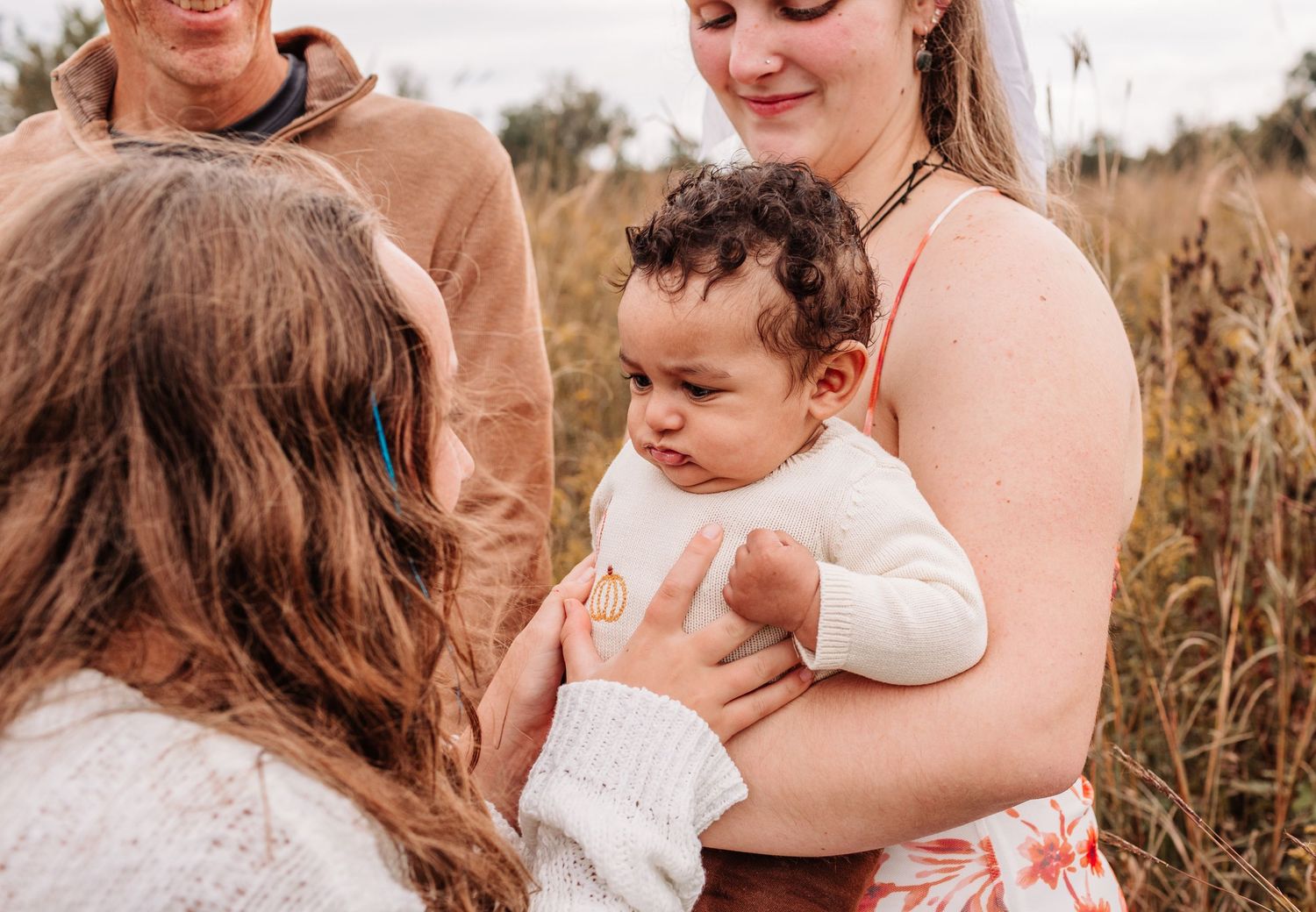 Woman holding baby in field while another person reaches out to touch the infant outdoors.