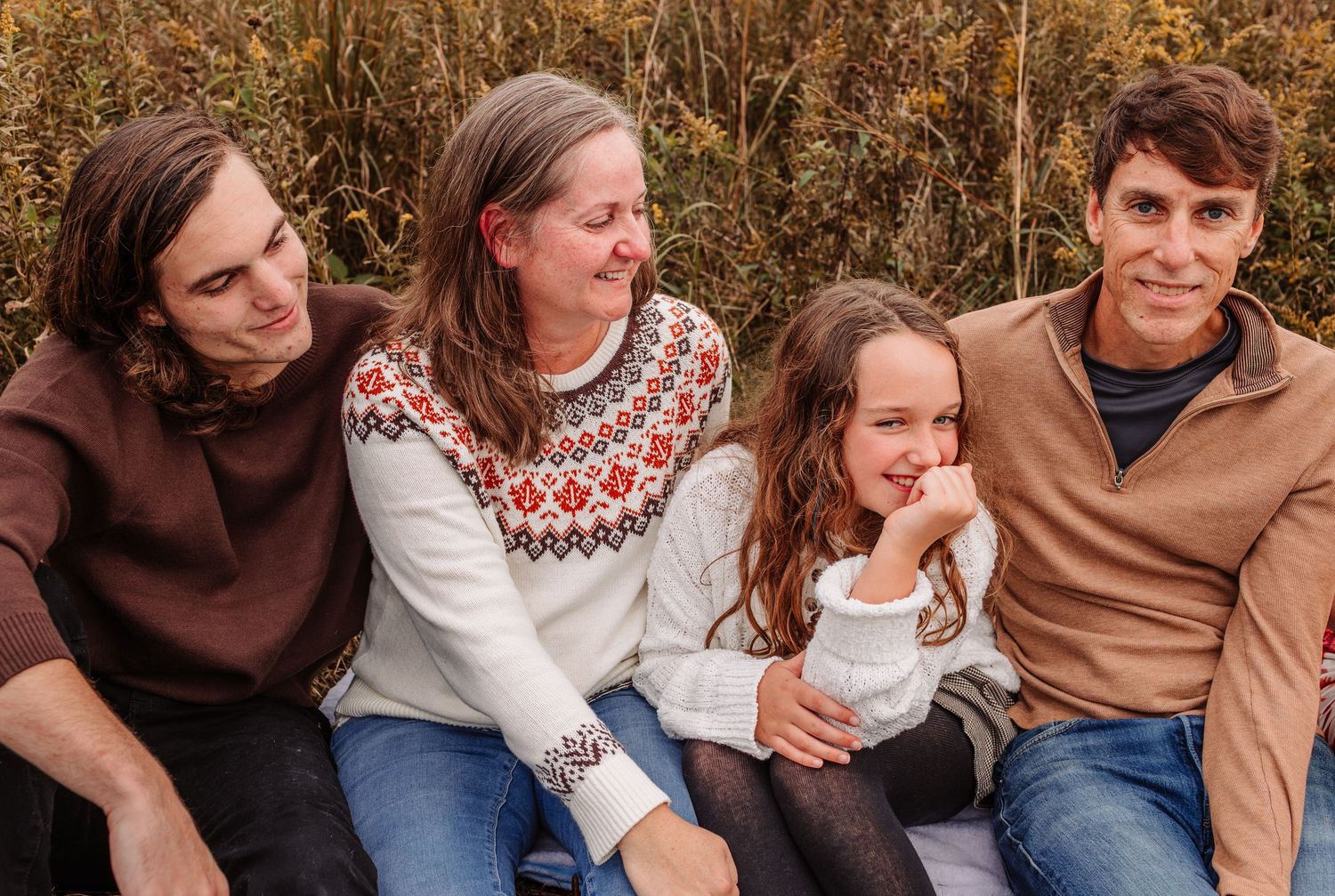 Family of four sitting outdoors in autumn, smiling warmly, wearing cozy sweaters amid golden foliage.